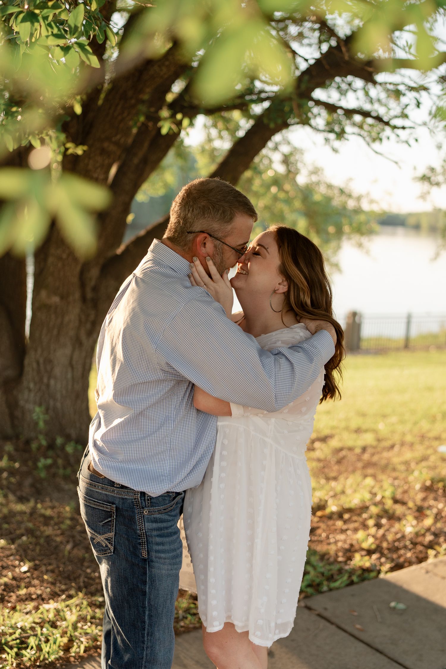 Intimate couple embrace during sunset photoshoot beneath leafy tree branches.