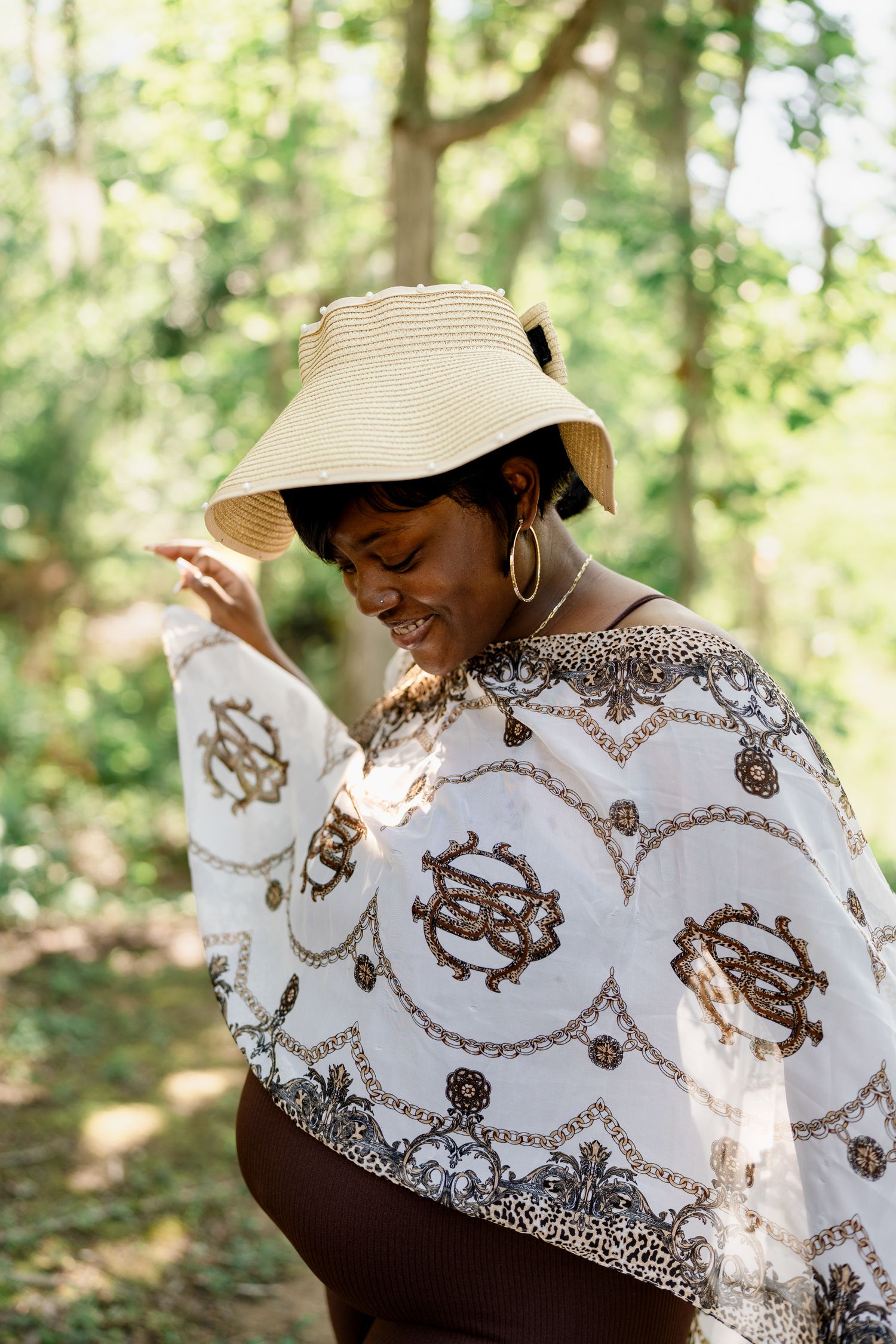 Person wearing straw hat and decorative white shawl smiles while adjusting their hat in dappled forest light.