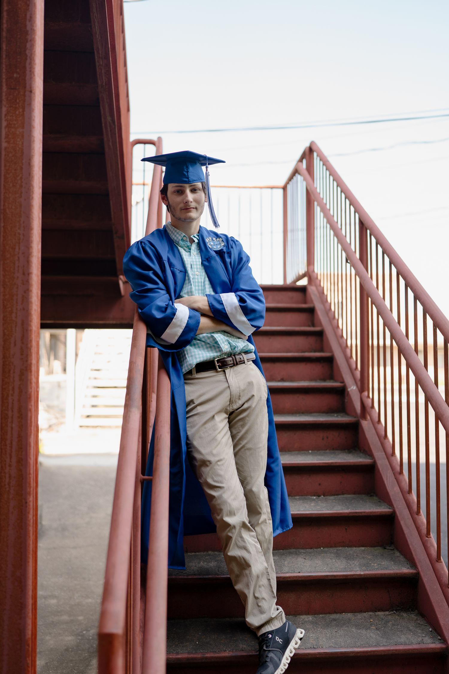 Graduate in blue cap and gown poses against red metal staircase during photo session.