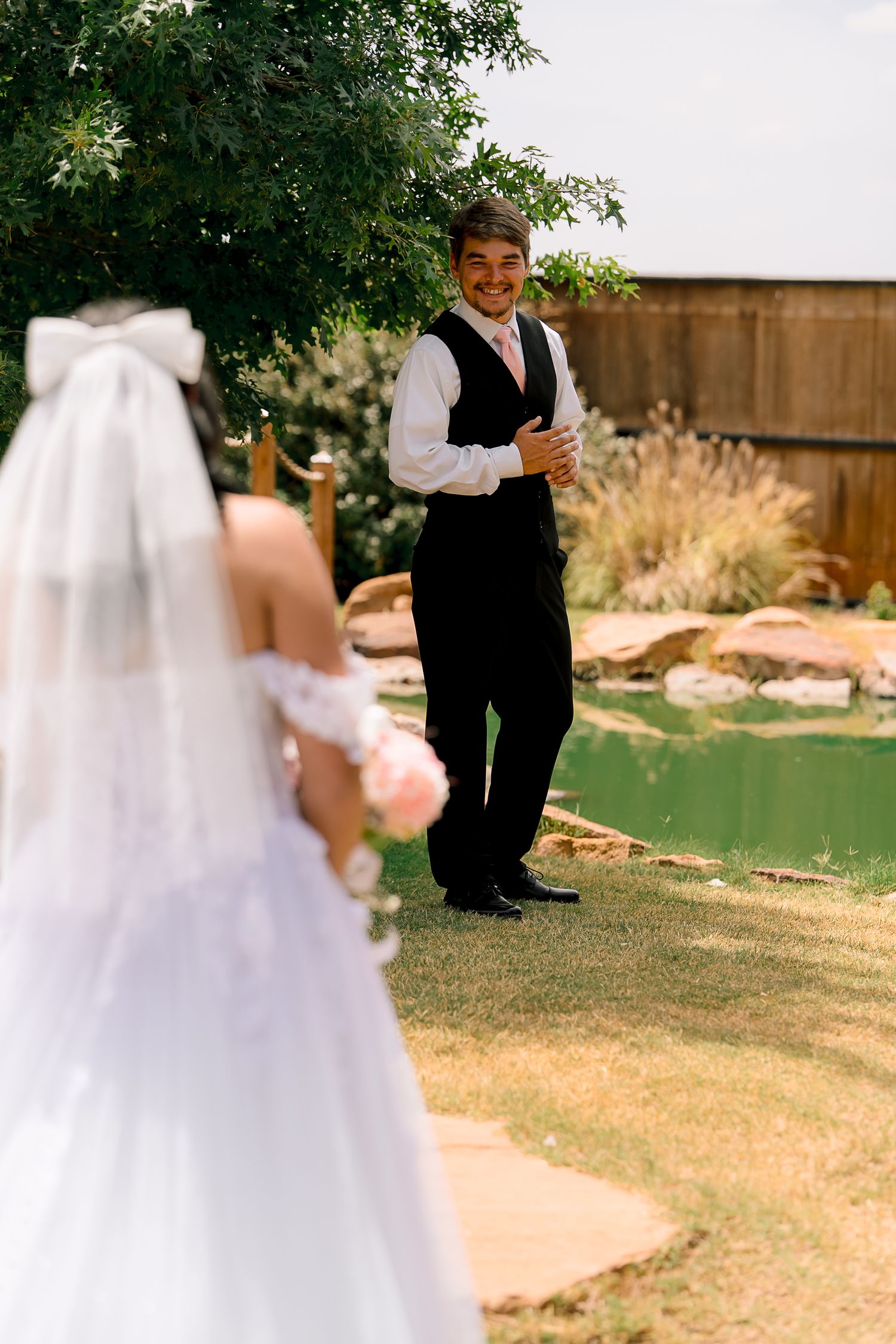 Groom smiling at bride during outdoor first look by a green pond.