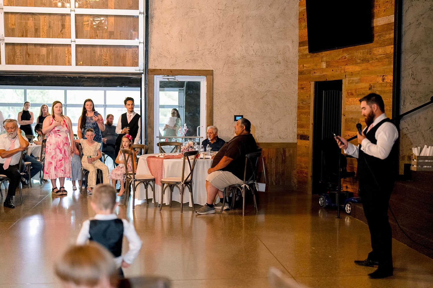 Wedding reception speech by groomsman in vest while guests sit at decorated tables in rustic venue.