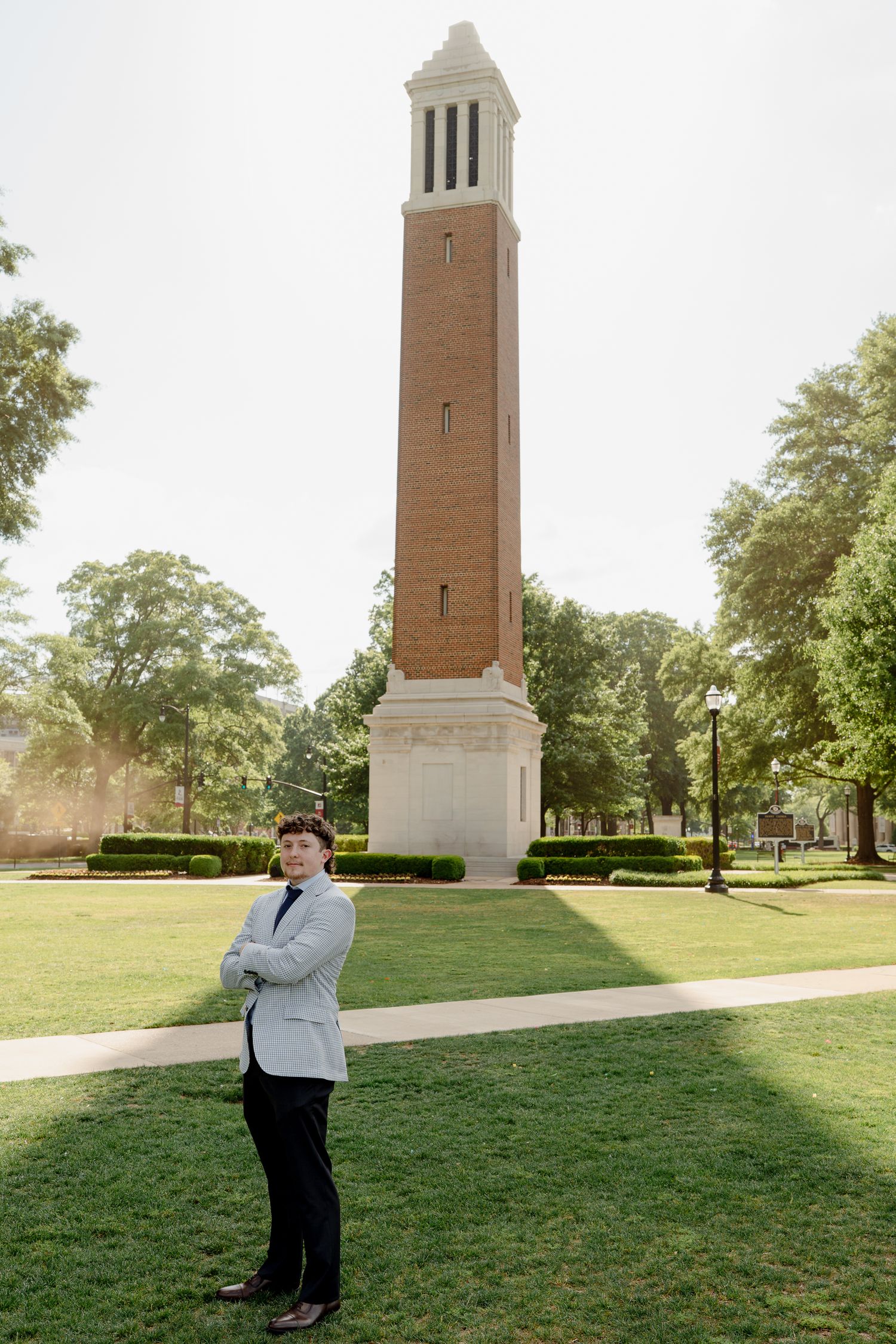 Graduate stands confidently with arms crossed before Denny Chimes bell tower at University of Alabama.