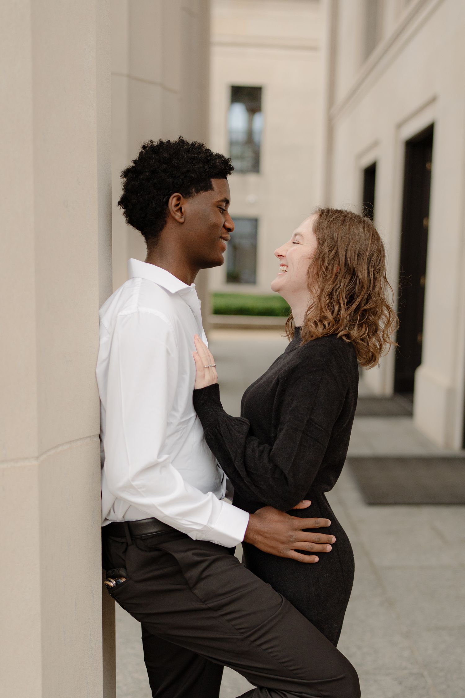 A young interracial couple shares a romantic moment, smiling at each other against a stone building column.