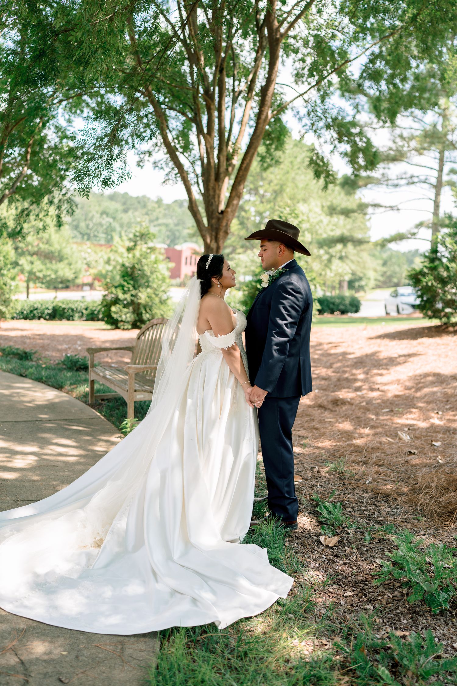Elegant wedding couple poses outdoors, bride in off-shoulder gown with long train and groom wearing western hat.