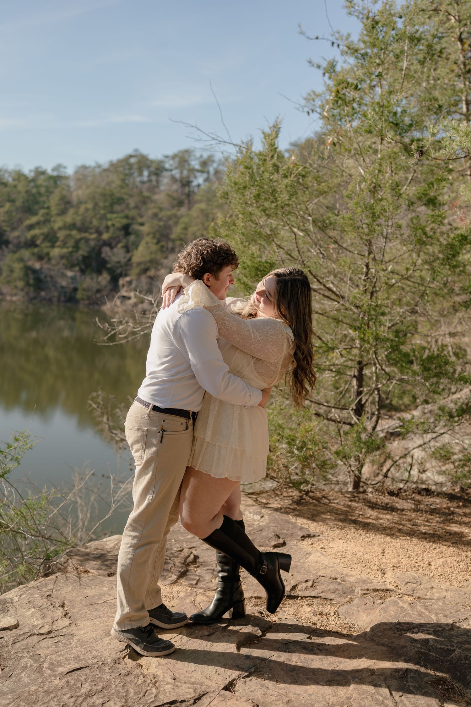 Couple embracing on rocky overlook above a serene lake surrounded by lush green trees on a sunny day.