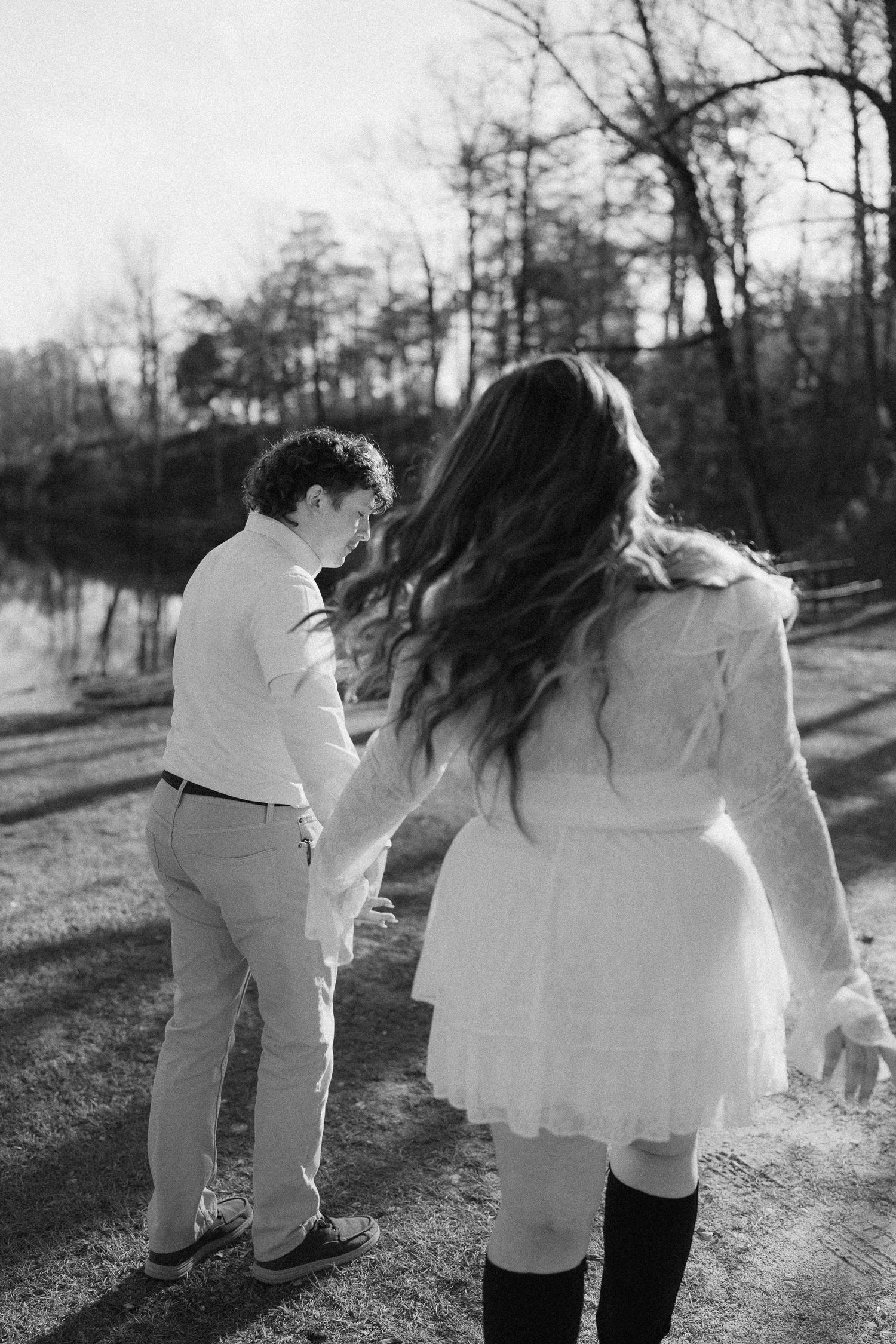 Black and white photo of couple holding hands walking near a sunlit lake with bare trees.