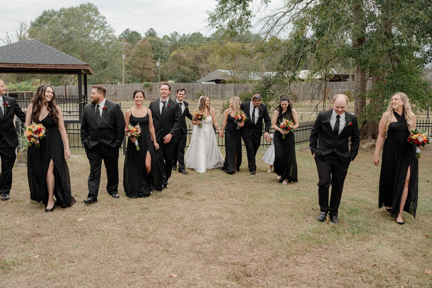 Wedding party members lined up outdoors wearing formal black attire with bride in white gown.