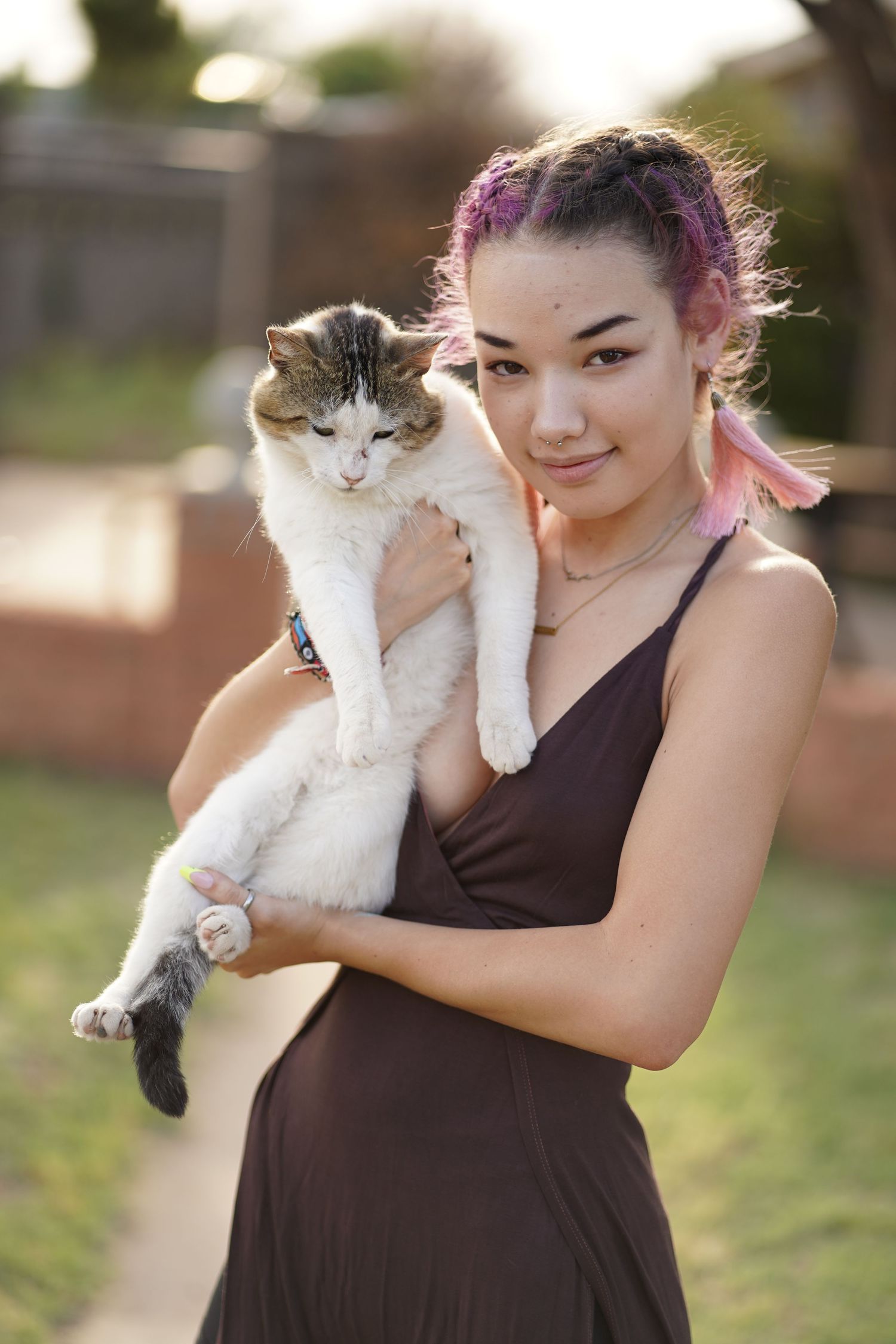 Person in a black dress holds a fluffy white and grey cat while standing outside.