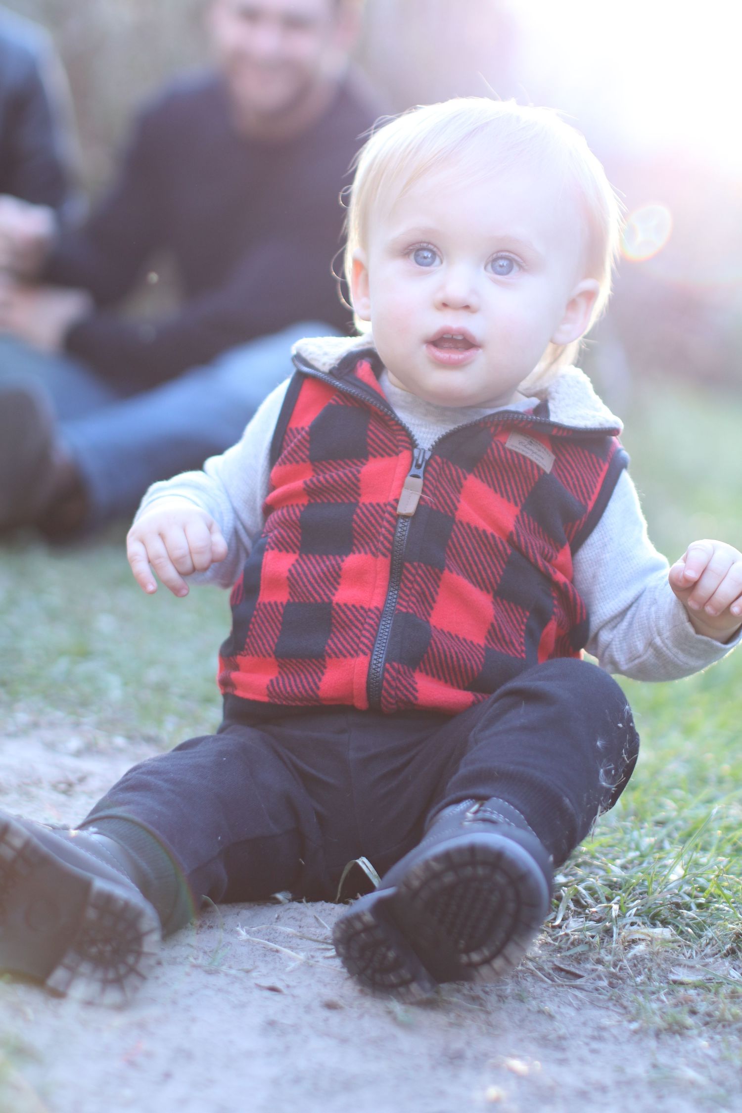 Toddler in red buffalo plaid vest sitting on ground with sunlit background.