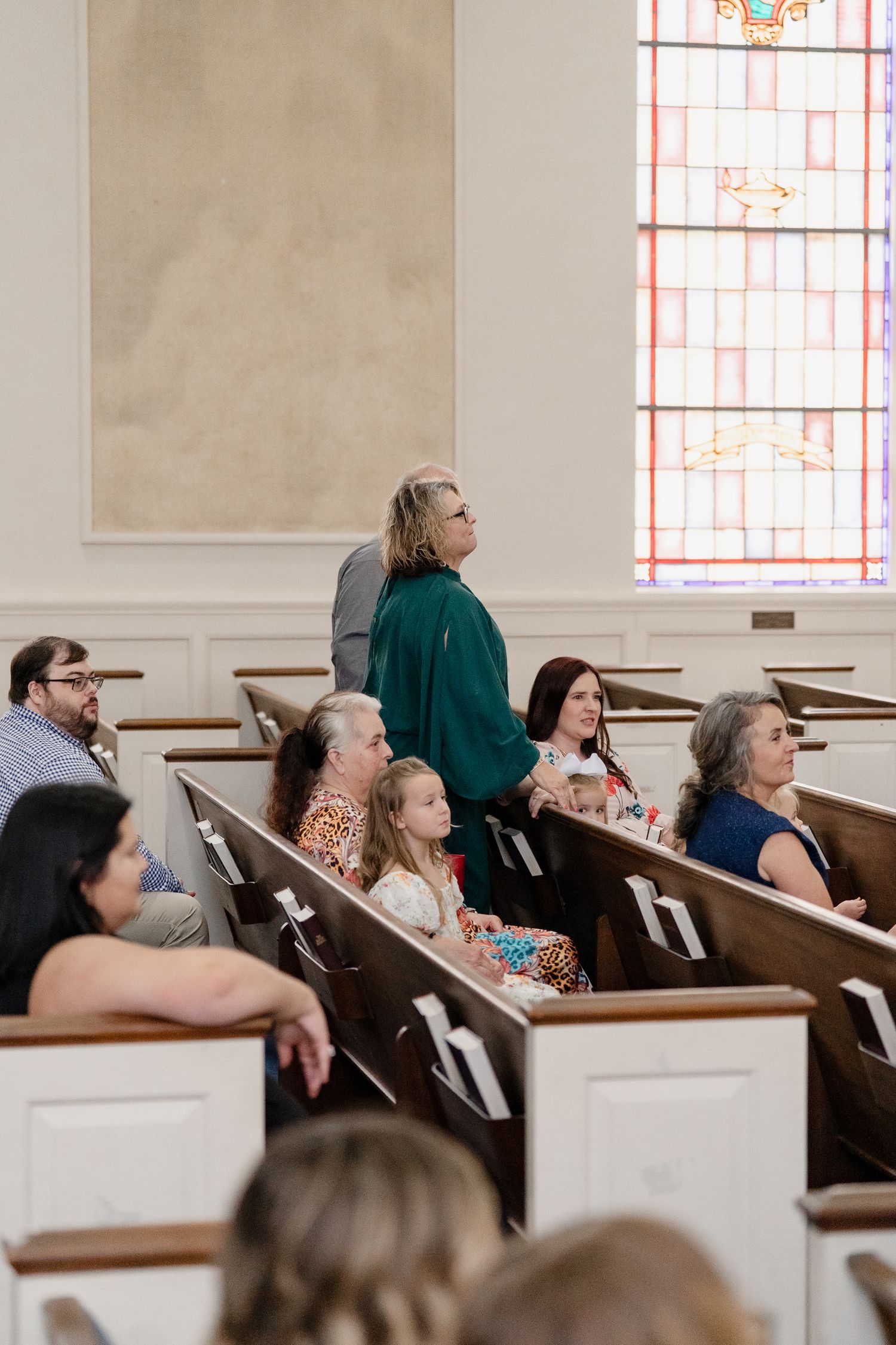 People seated in traditional wooden church pews with stained glass windows visible in the background during a service.