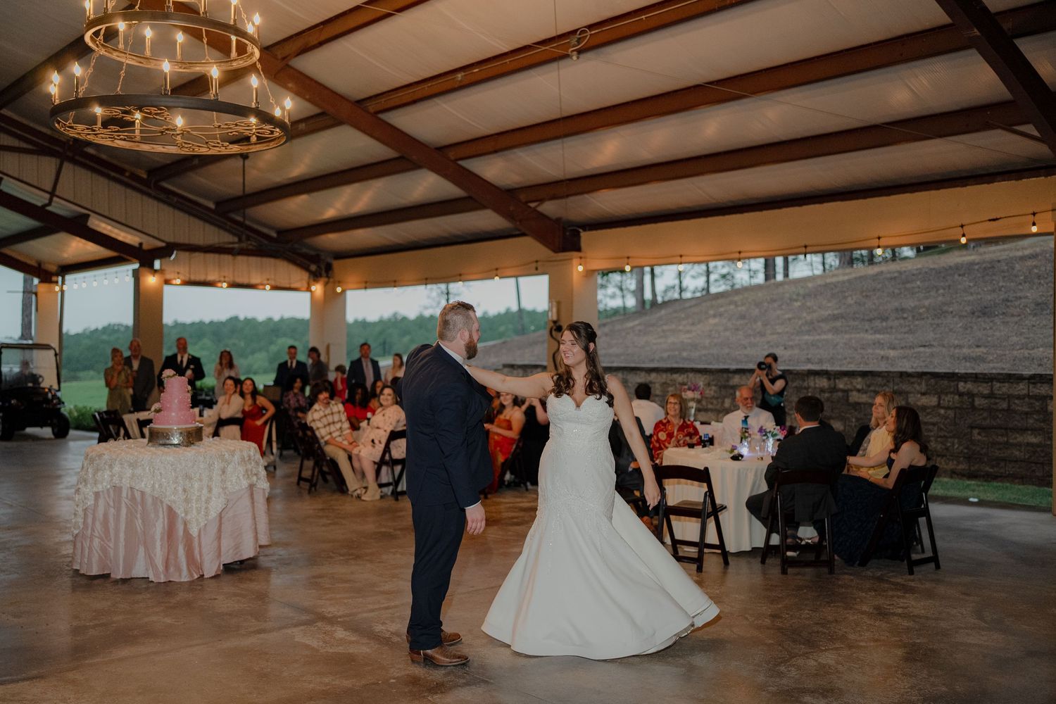 Bride and groom share first dance in rustic open-air reception venue with string lights and guests.