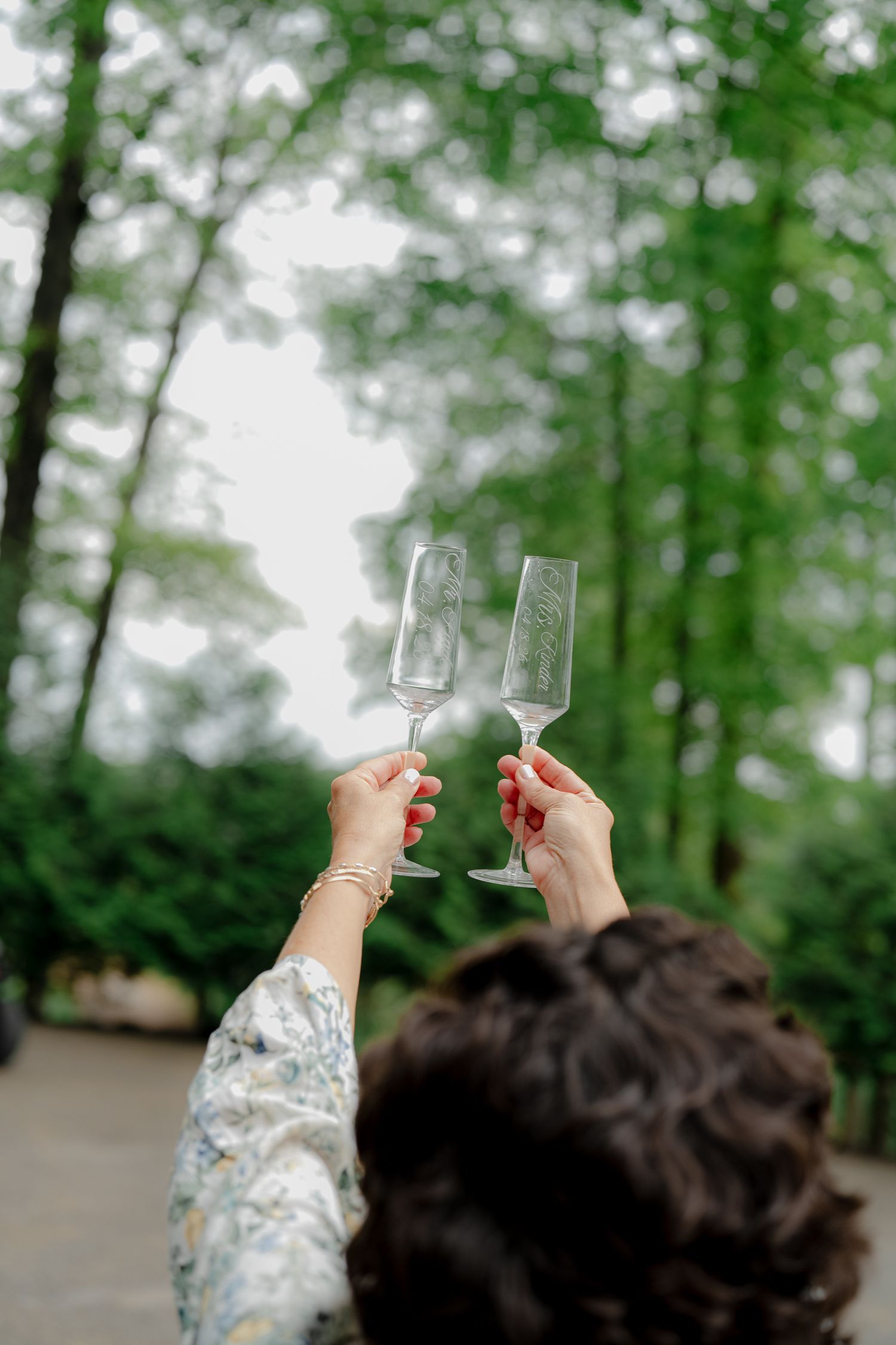 Person raising two empty champagne flutes in celebration outdoors surrounded by lush green trees.