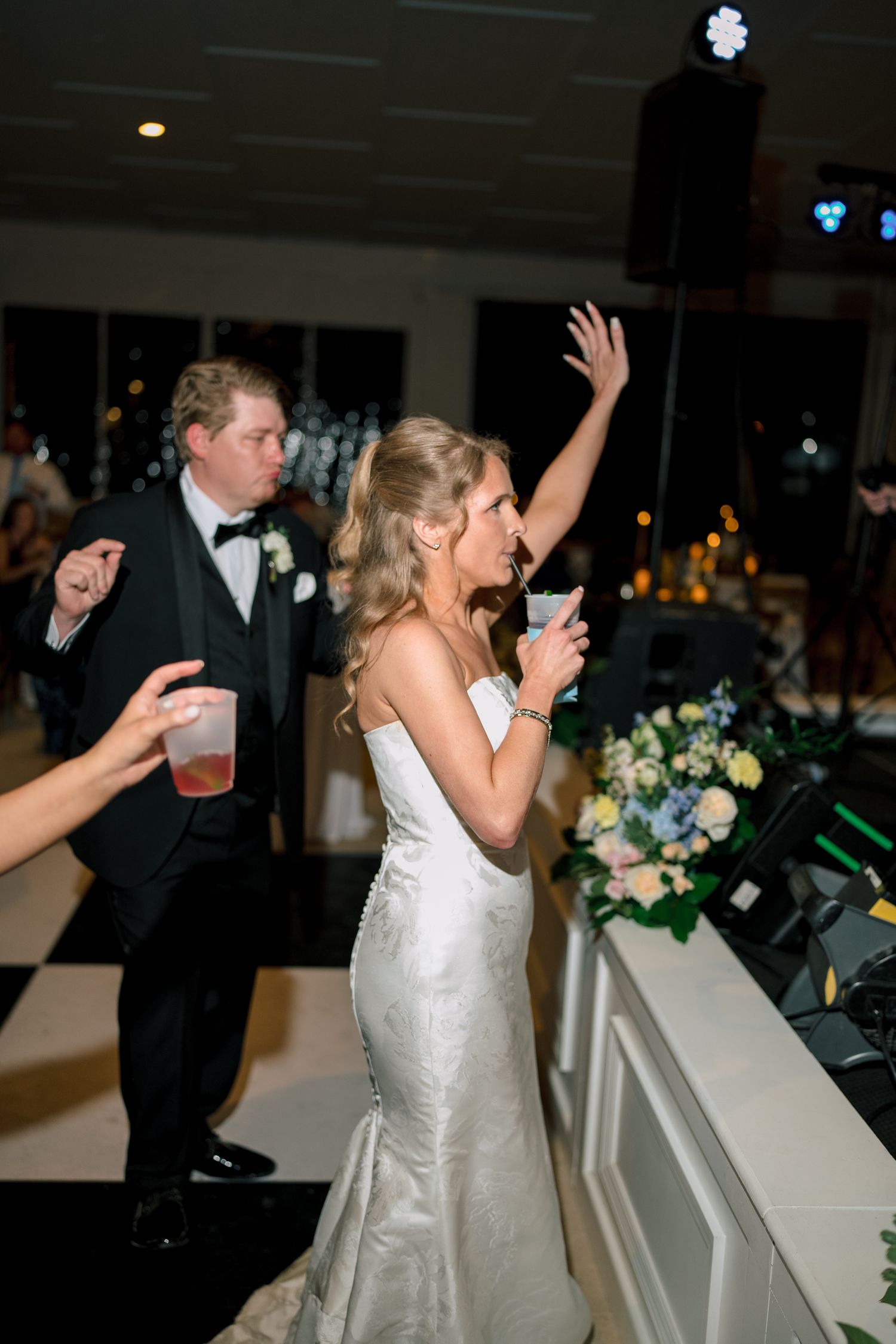 Bride in white gown raises hand while dancing at wedding reception, groom in tuxedo behind her.
