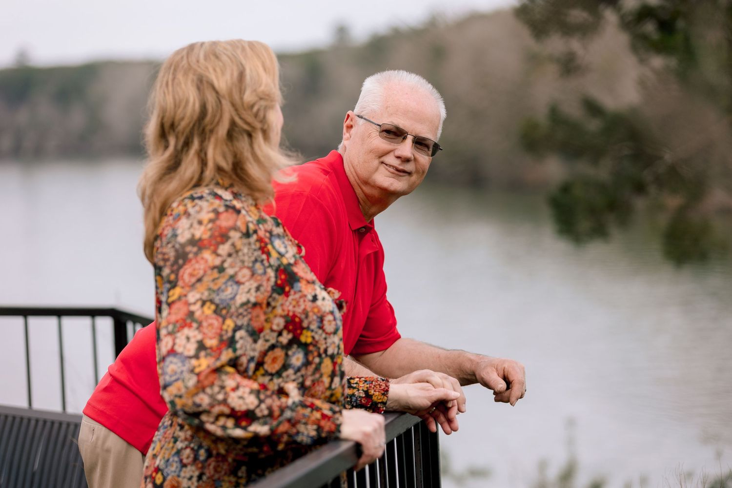 Older couple holding hands on a railing overlooking a serene lake, man in red polo shirt.