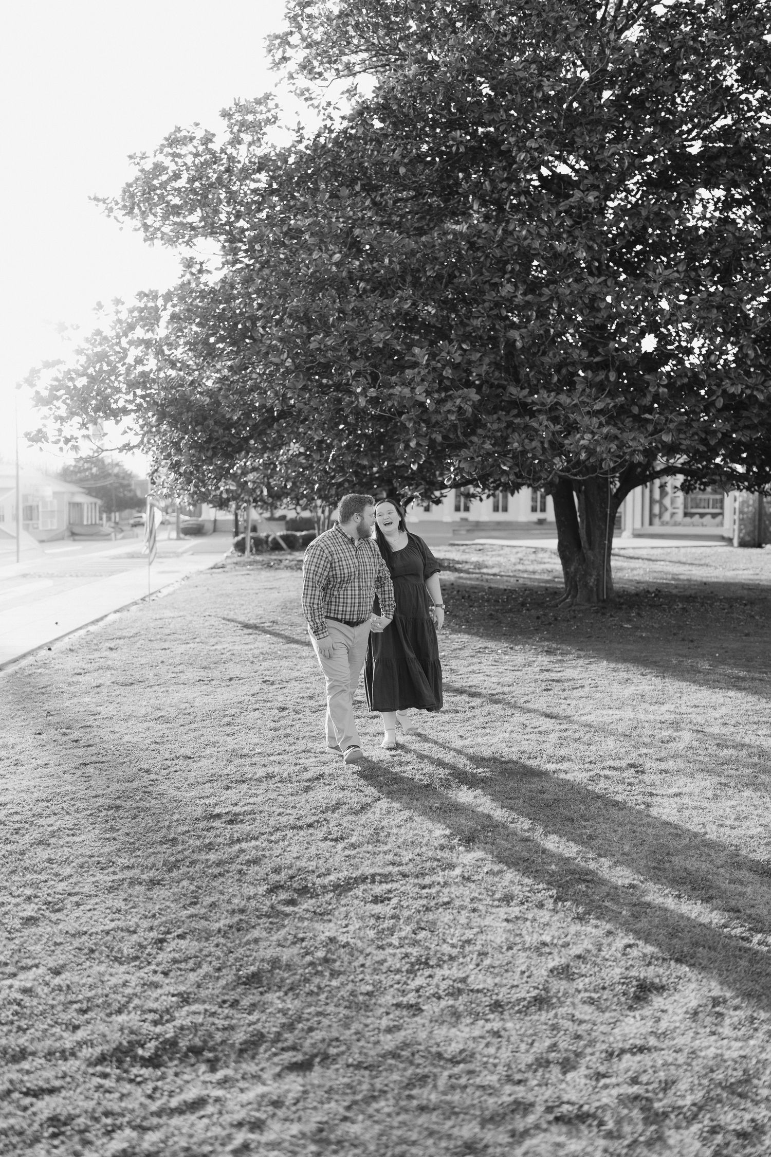 Black and white photo of a couple walking together on grass near a large tree in a sunny park.