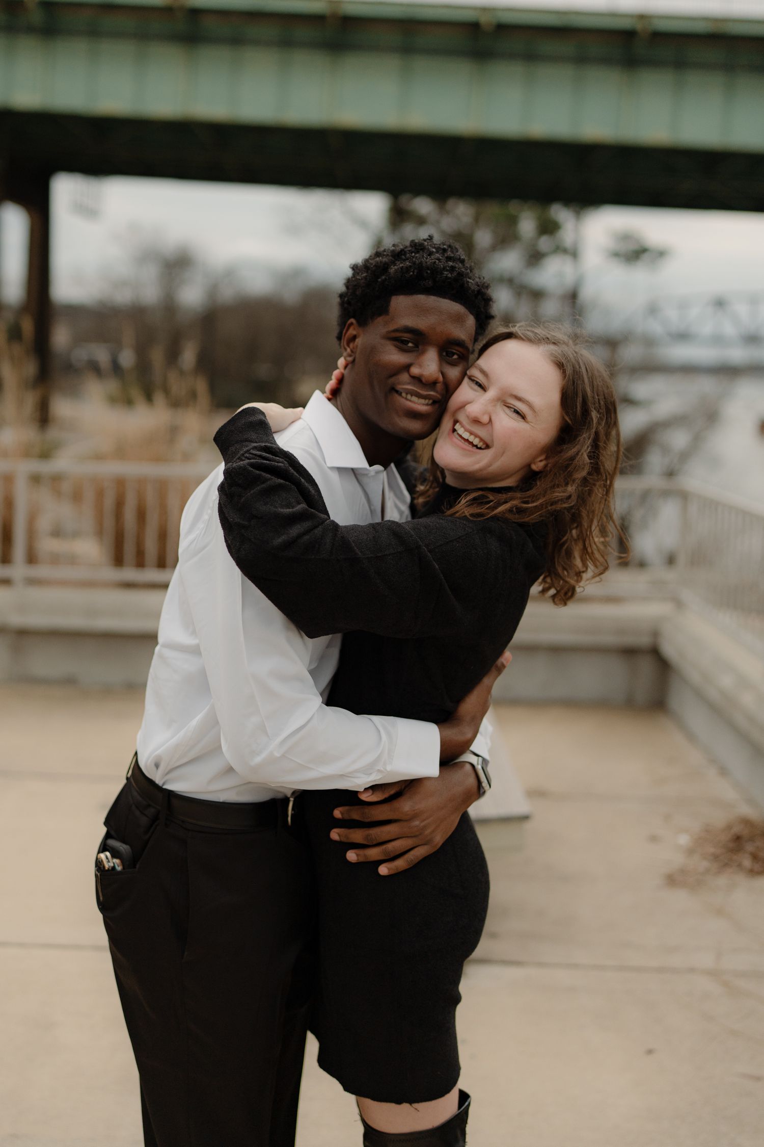 A joyful interracial couple embracing and smiling outdoors near a bridge, dressed in black and white formal attire.