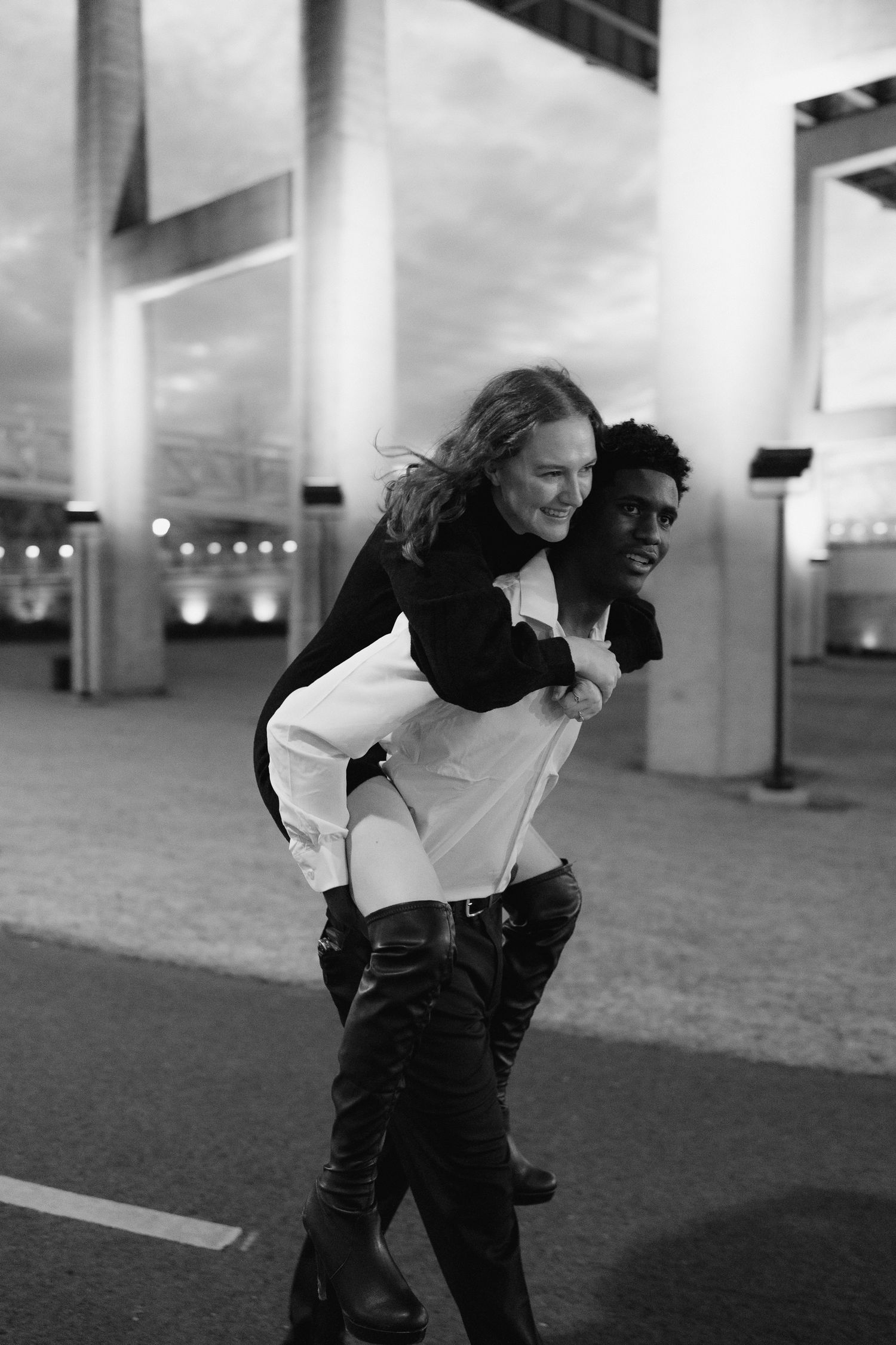 Black and white photo of a laughing woman getting a piggyback ride from her partner under a bridge.