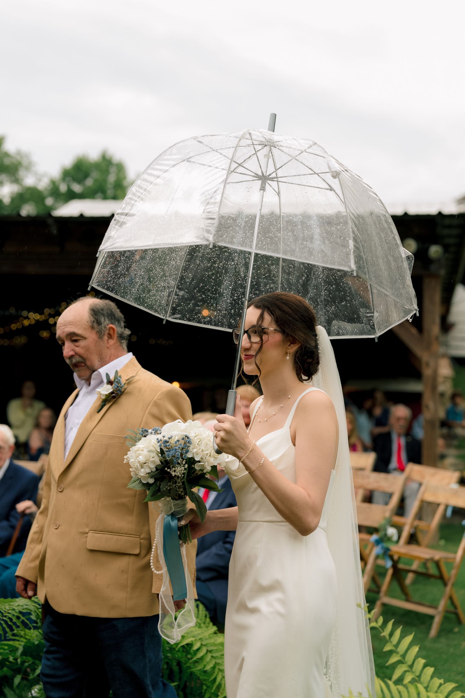 Bride walking down outdoor aisle with father under clear umbrella in the rain holding white bouquet.