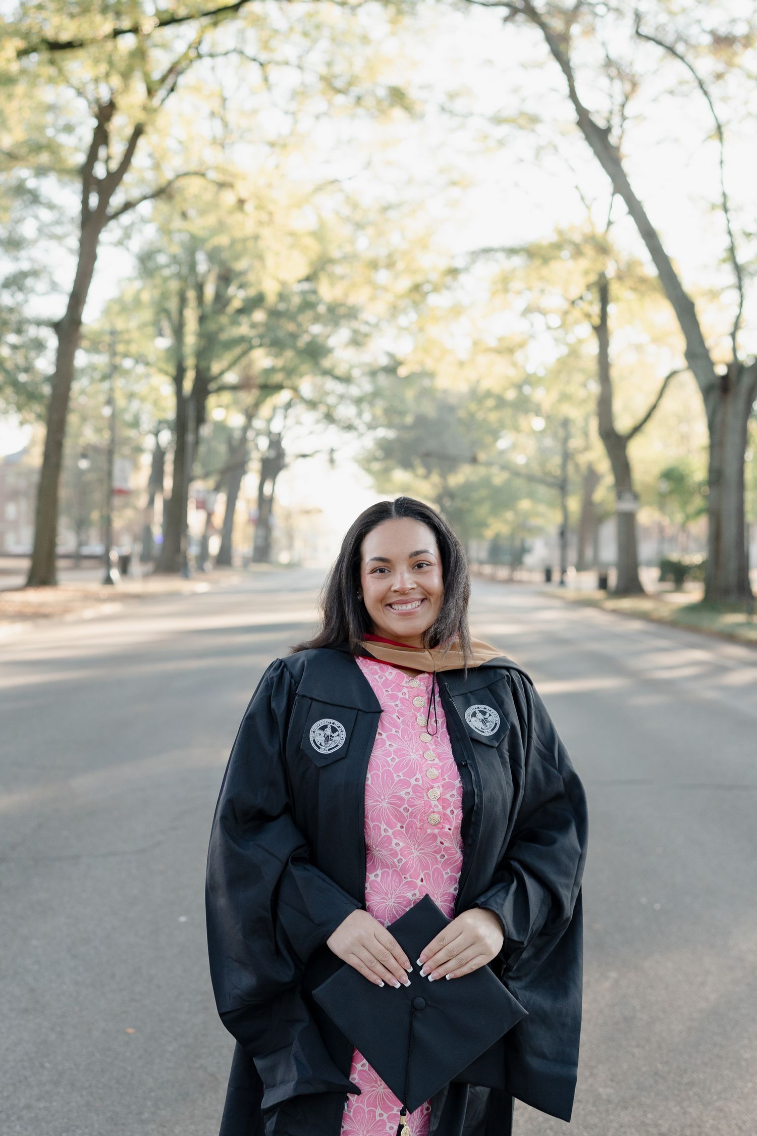 A graduate in academic regalia and pink dress holds a diploma on a tree-lined campus street.
