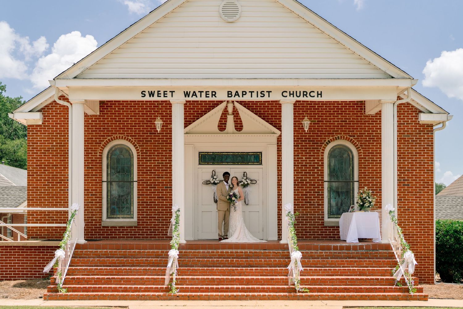 Bride and groom pose at Sweet Water Baptist Church entrance, a classic red brick building with white columns.