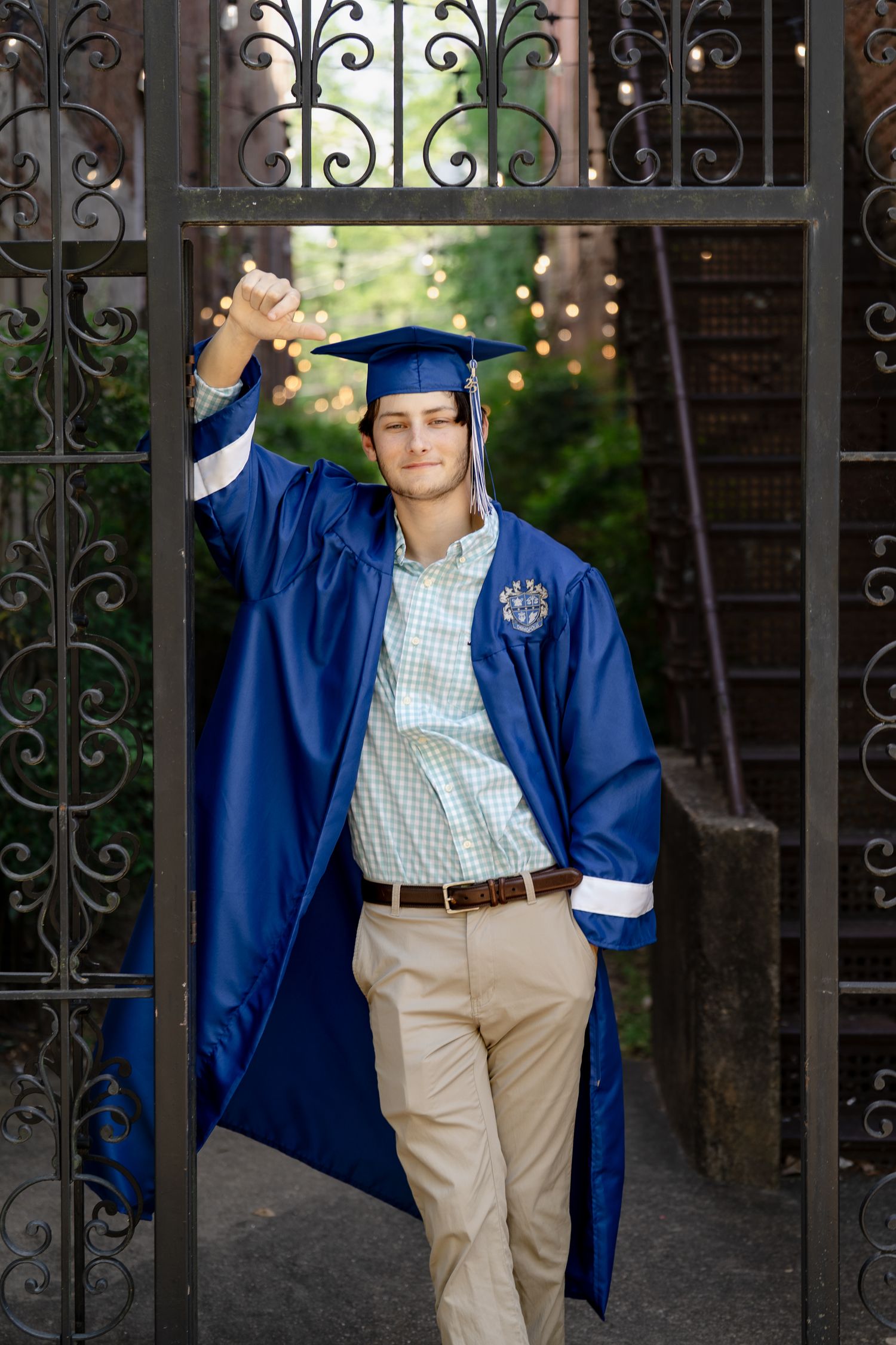 Graduate poses playfully in blue cap and gown by ornate iron gate.