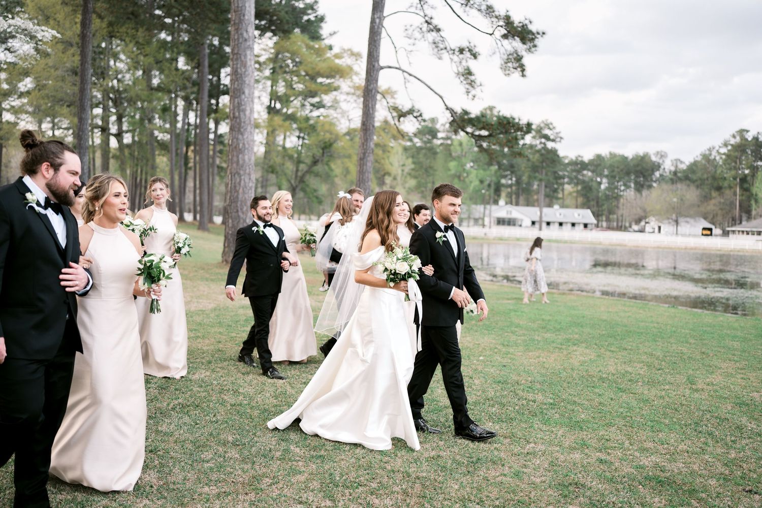 Wedding party walks together across lawn with pine trees and water in background.