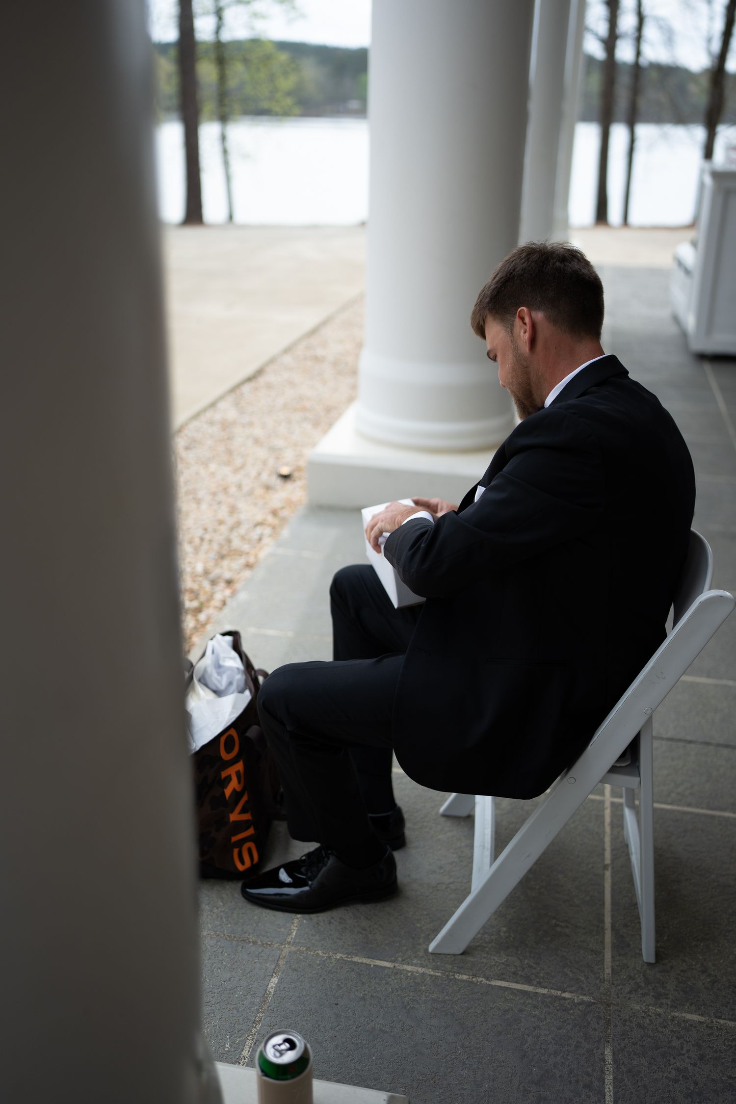Groom in black suit sits alone on porch chair overlooking a lake.