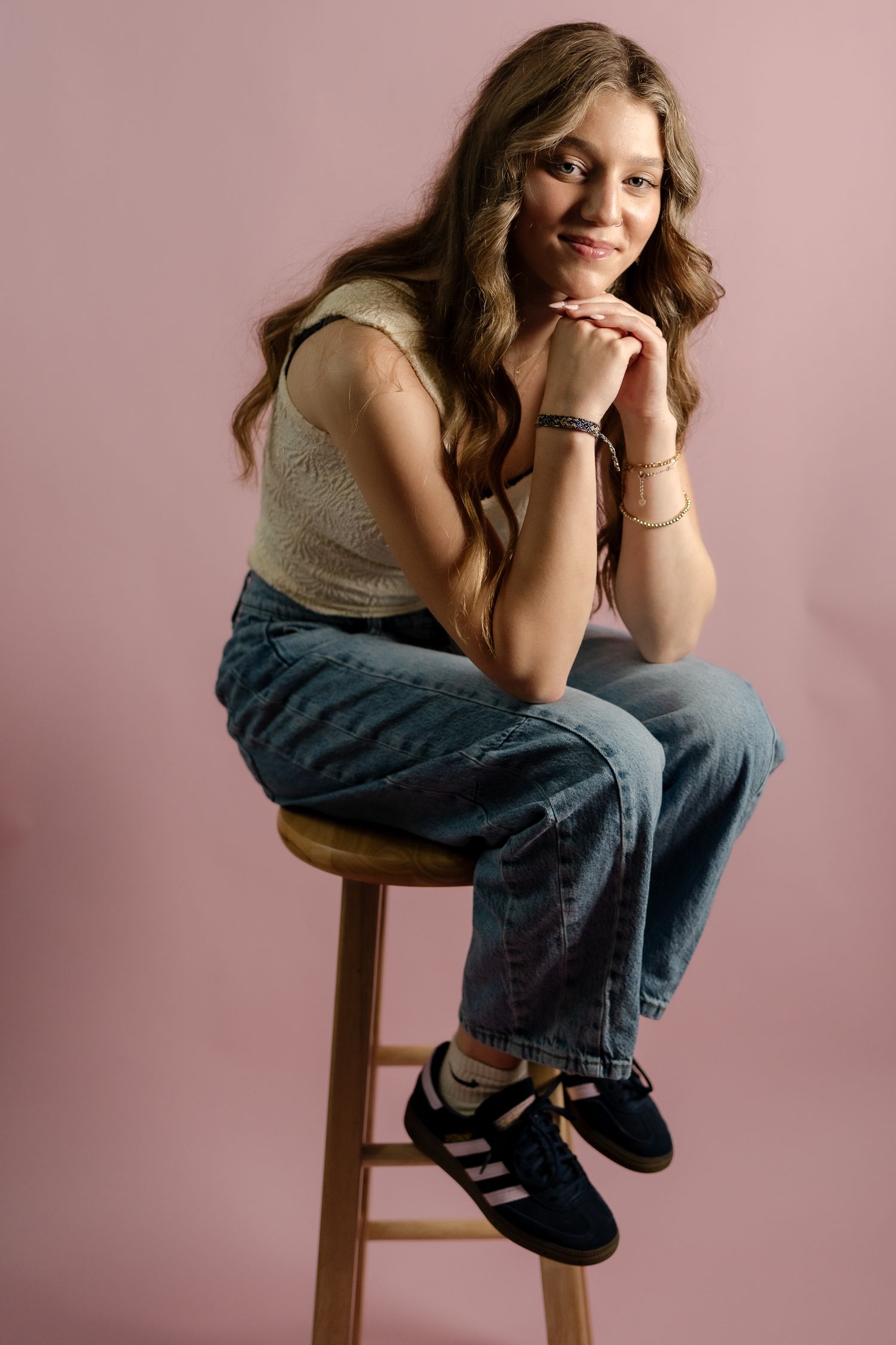 Stylish portrait of woman in 90s inspired outfit sitting on wooden stool against pink background.