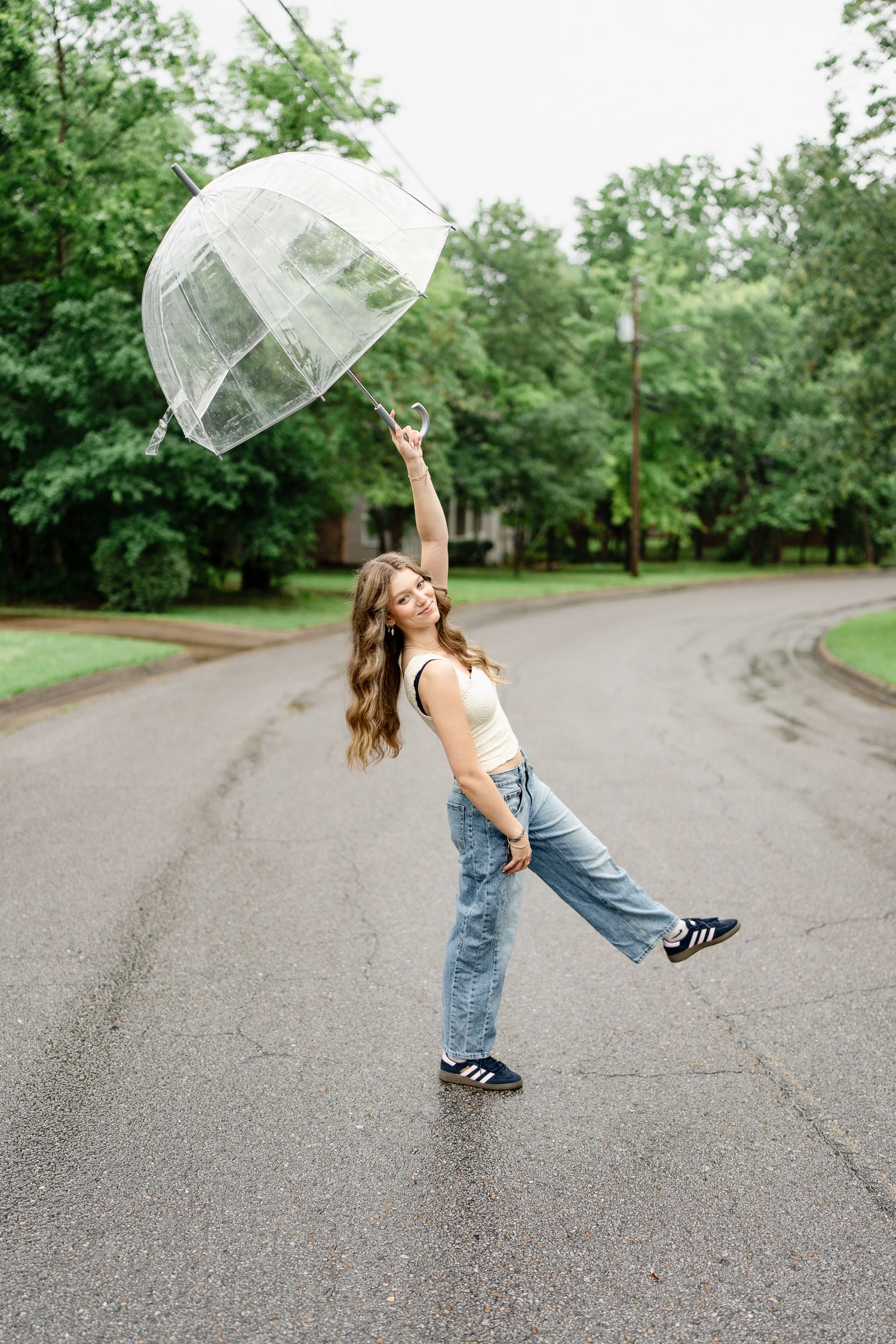 A playful scene on a tree-lined road with someone twirling a clear umbrella on an overcast day.