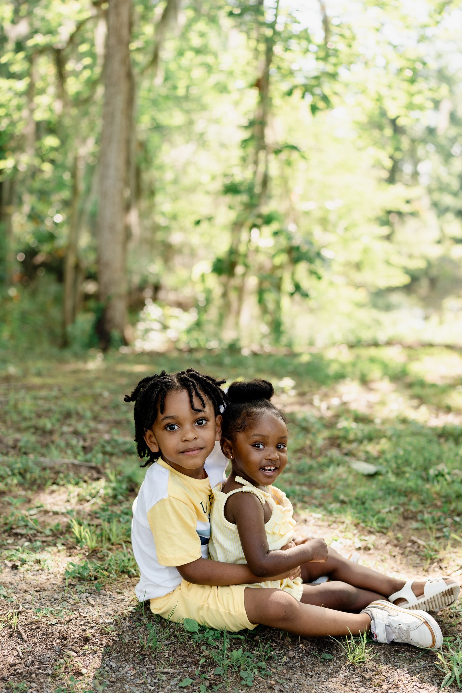Two children in casual attire sitting together on forest ground.