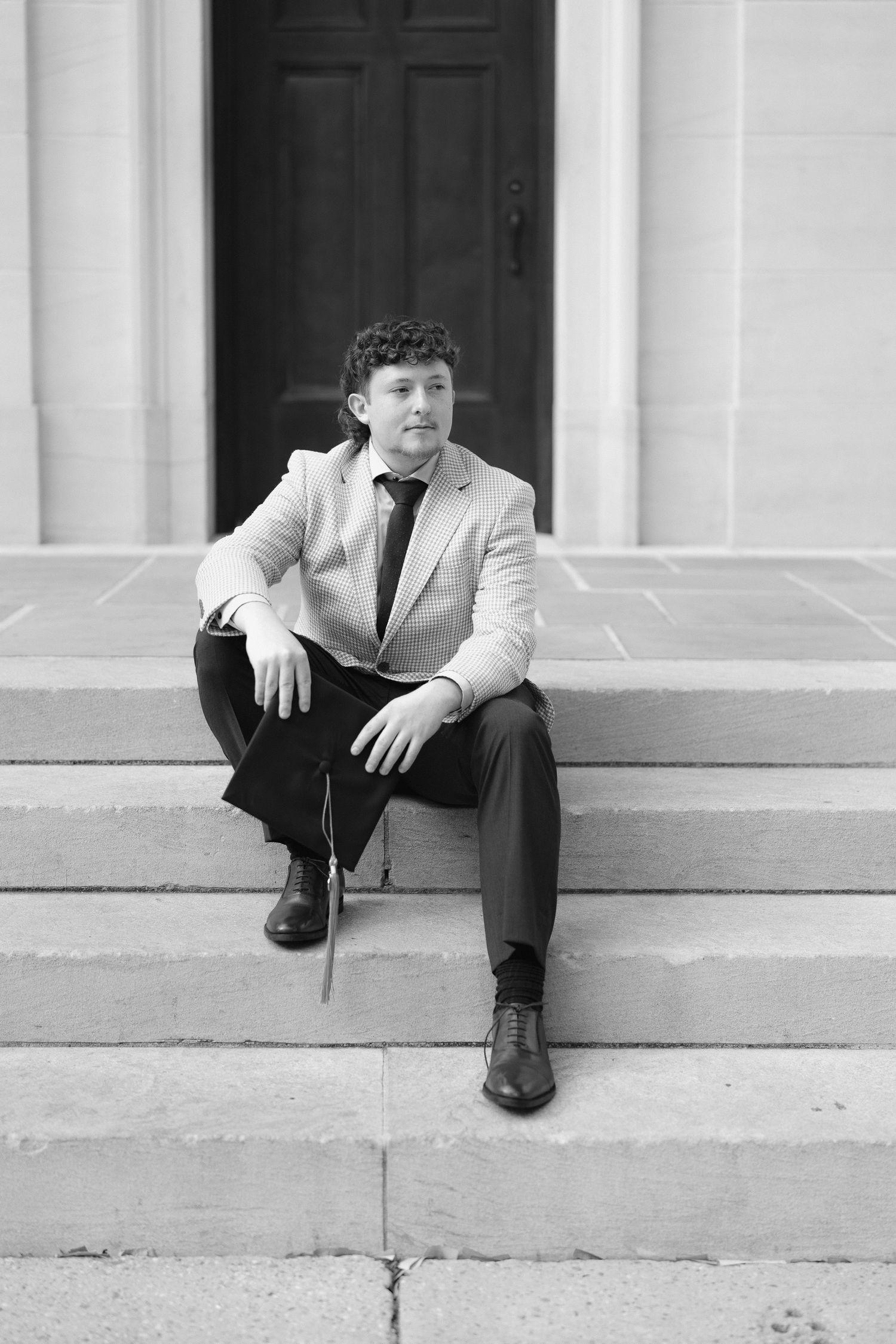 Black and white portrait of young man in blazer and tie sitting on stone steps outdoors.