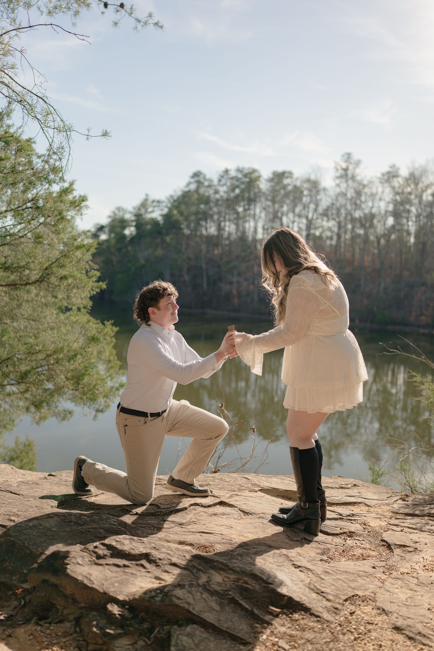 Man on one knee proposing to woman in cream dress on rocky overlook beside peaceful lake.