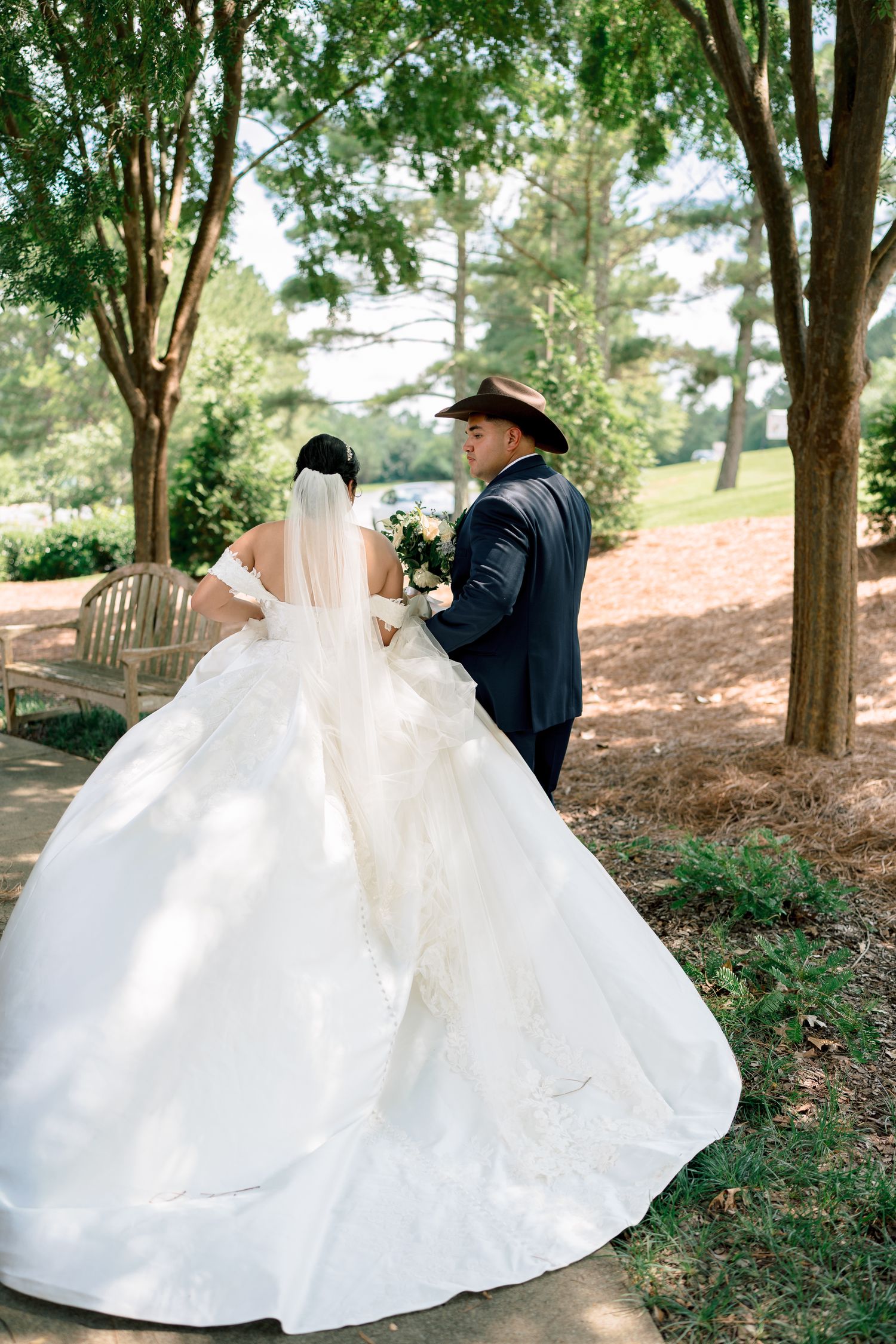 A stunning wedding photo showing a bride in a flowing white ballgown and groom in navy suit with cowboy hat under trees.