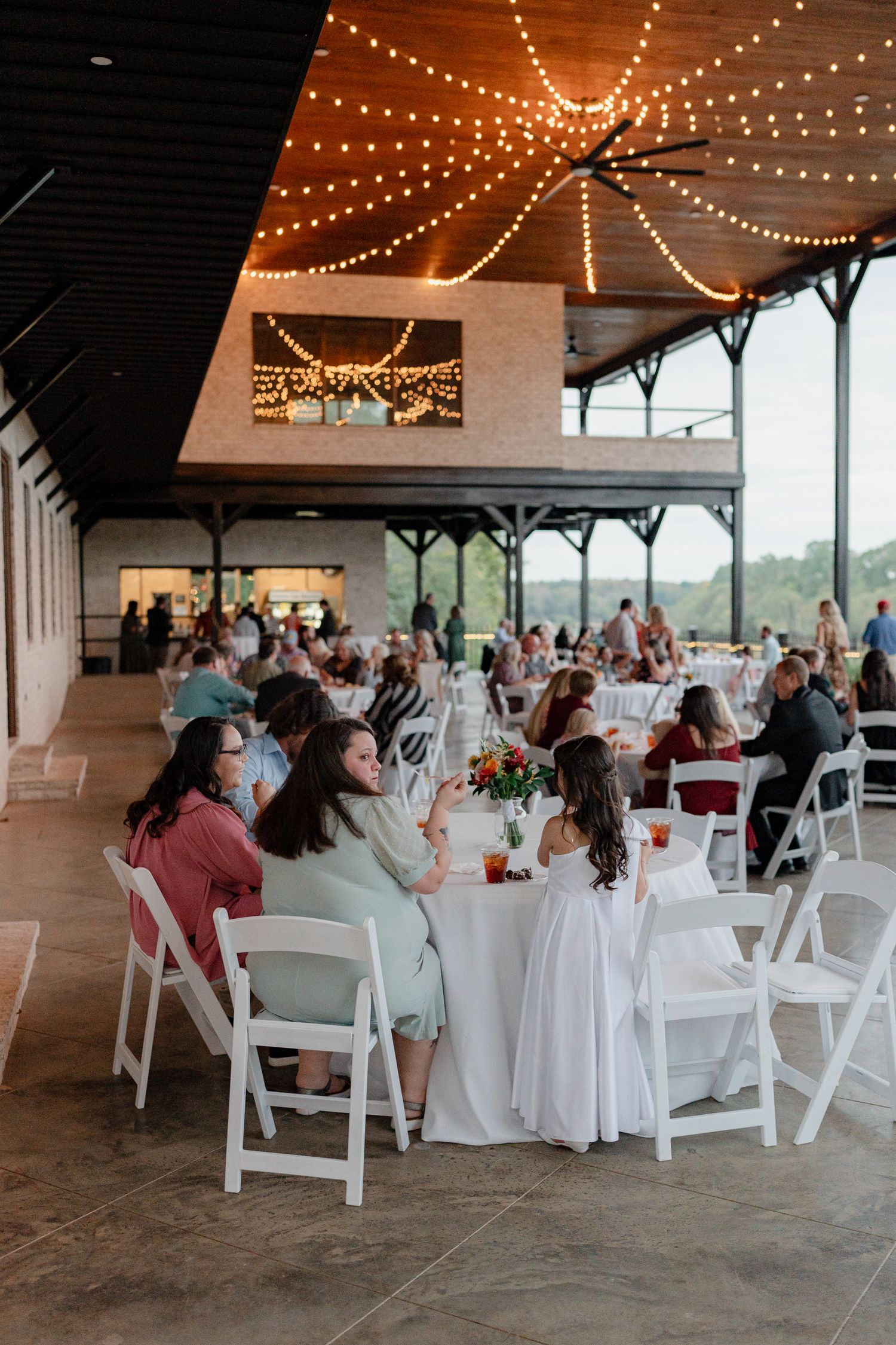 Wedding reception setup under string lights with white tables and chairs in an open-air venue.