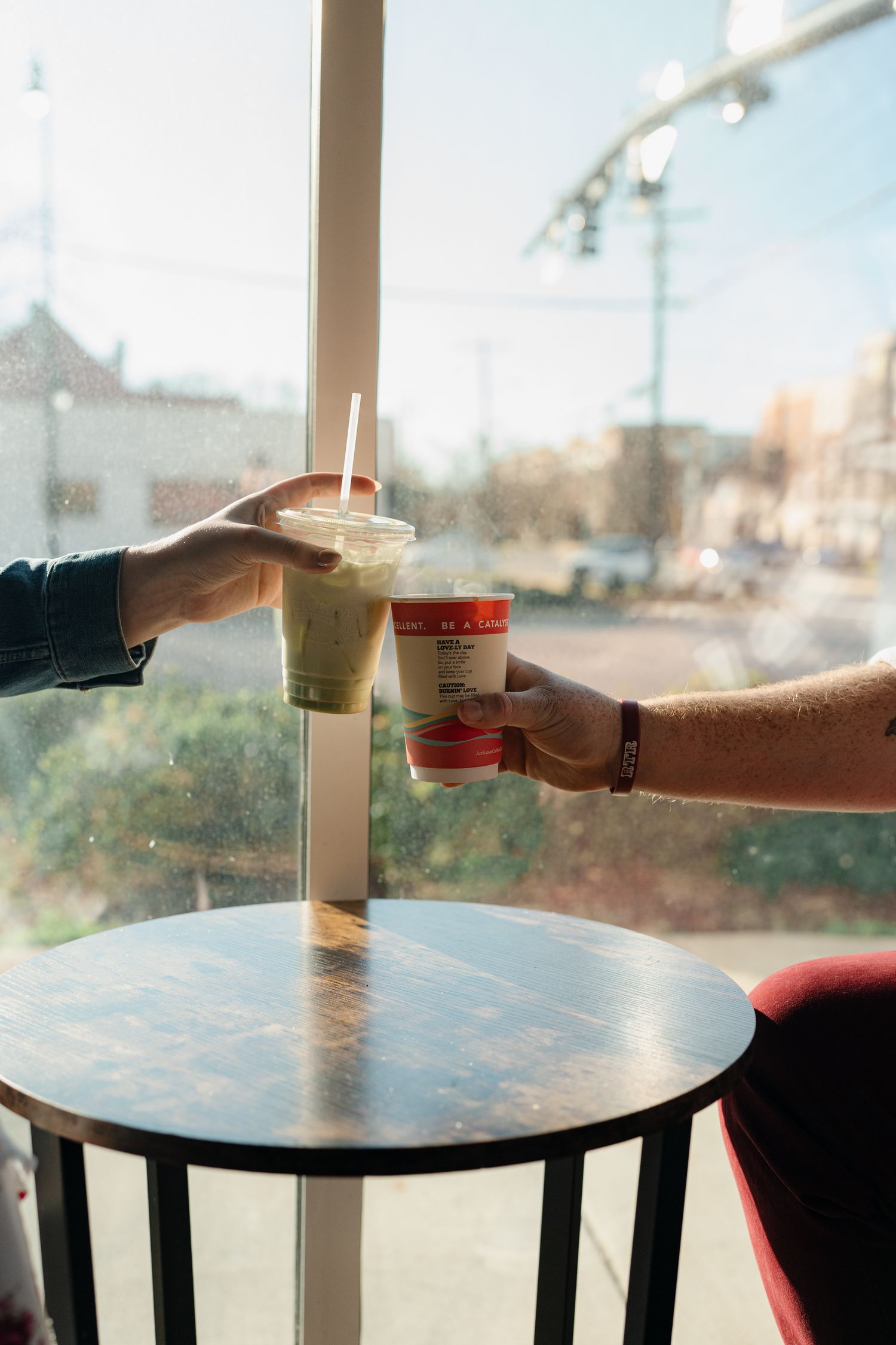 A hand reaches for a beverage on a round table by a window with sunlight streaming in.