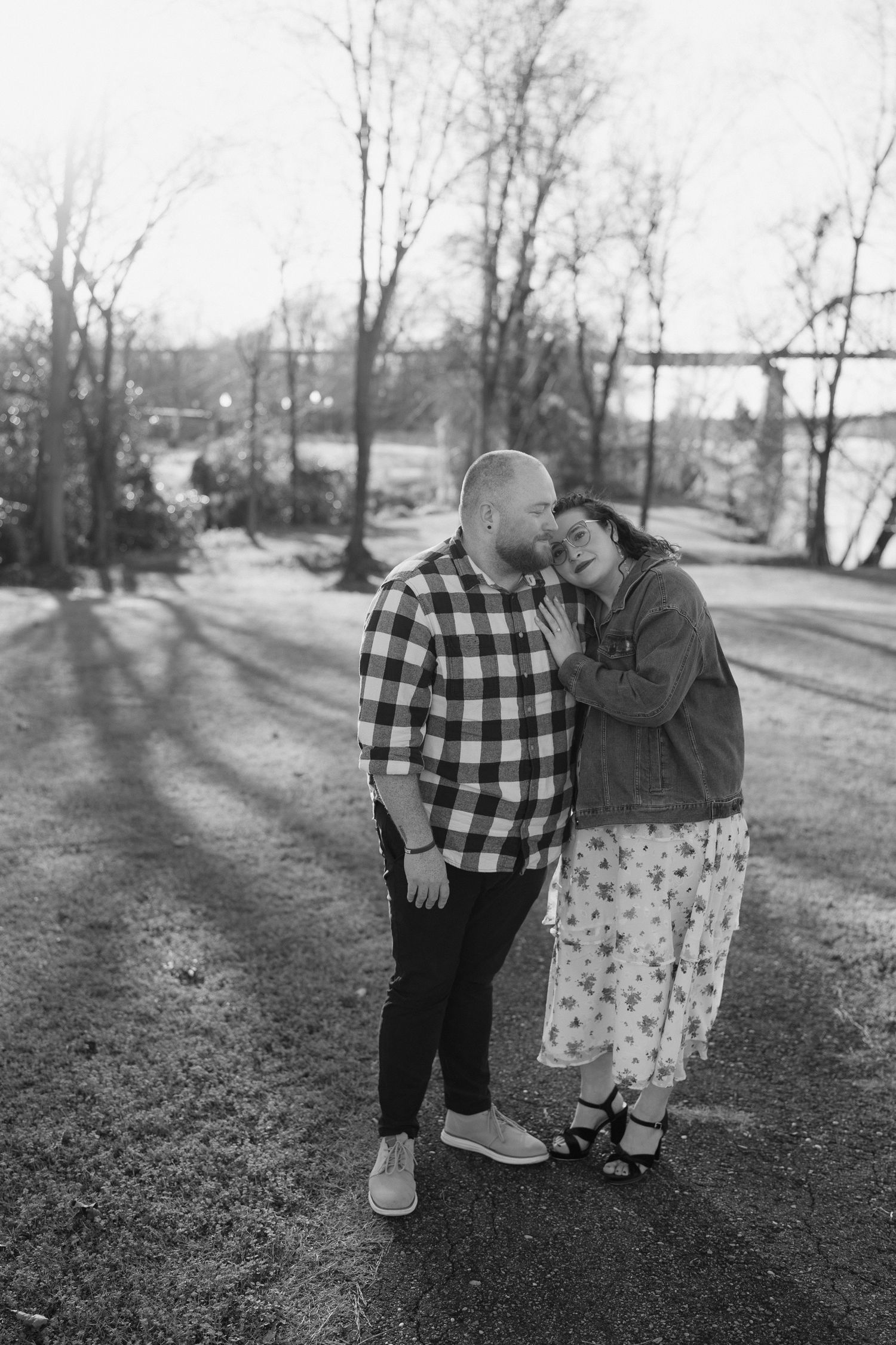 A black and white photo of a couple in casual attire standing together on a path with bare trees in background.