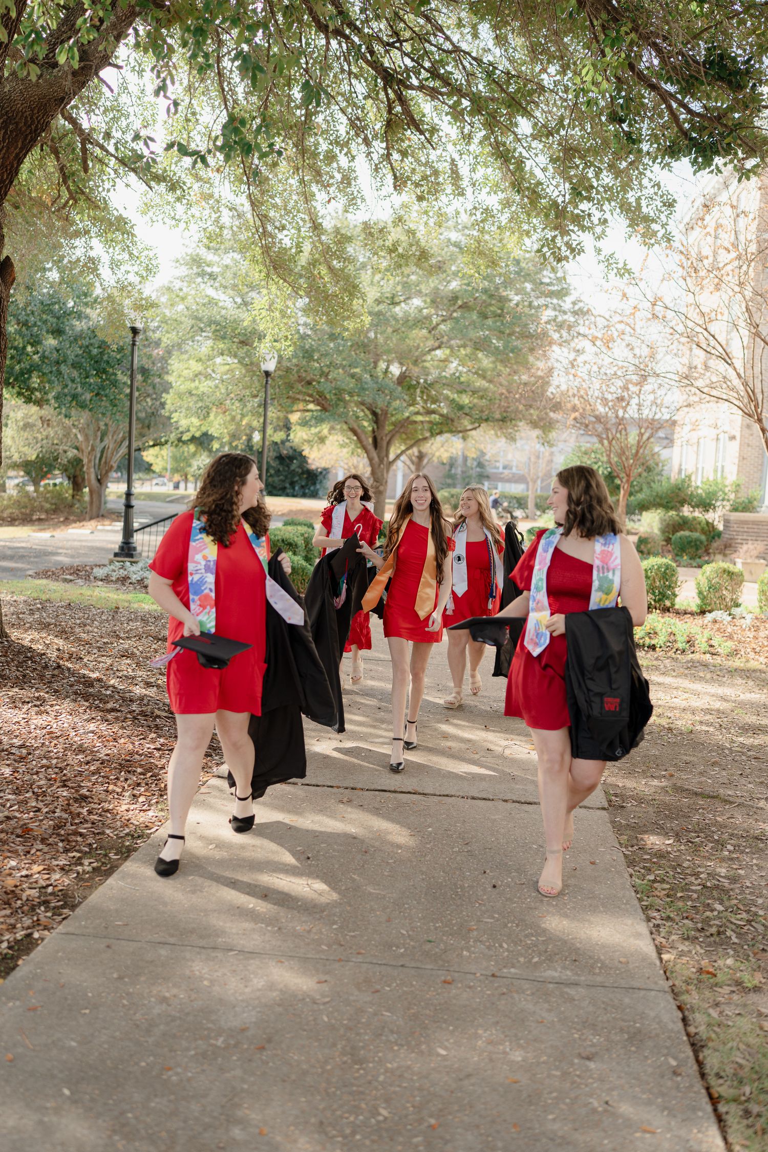 A group of dancers in red and black outfits perform synchronized poses while walking along a tree-lined path.