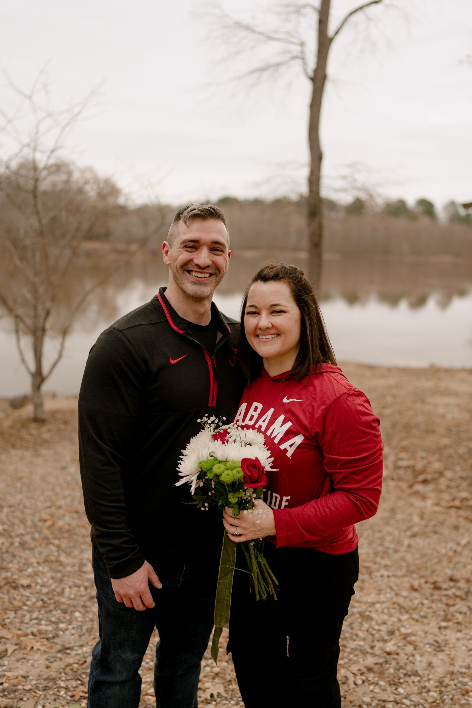 A couple in Alabama sports gear poses with flowers by a serene lake on an overcast day.