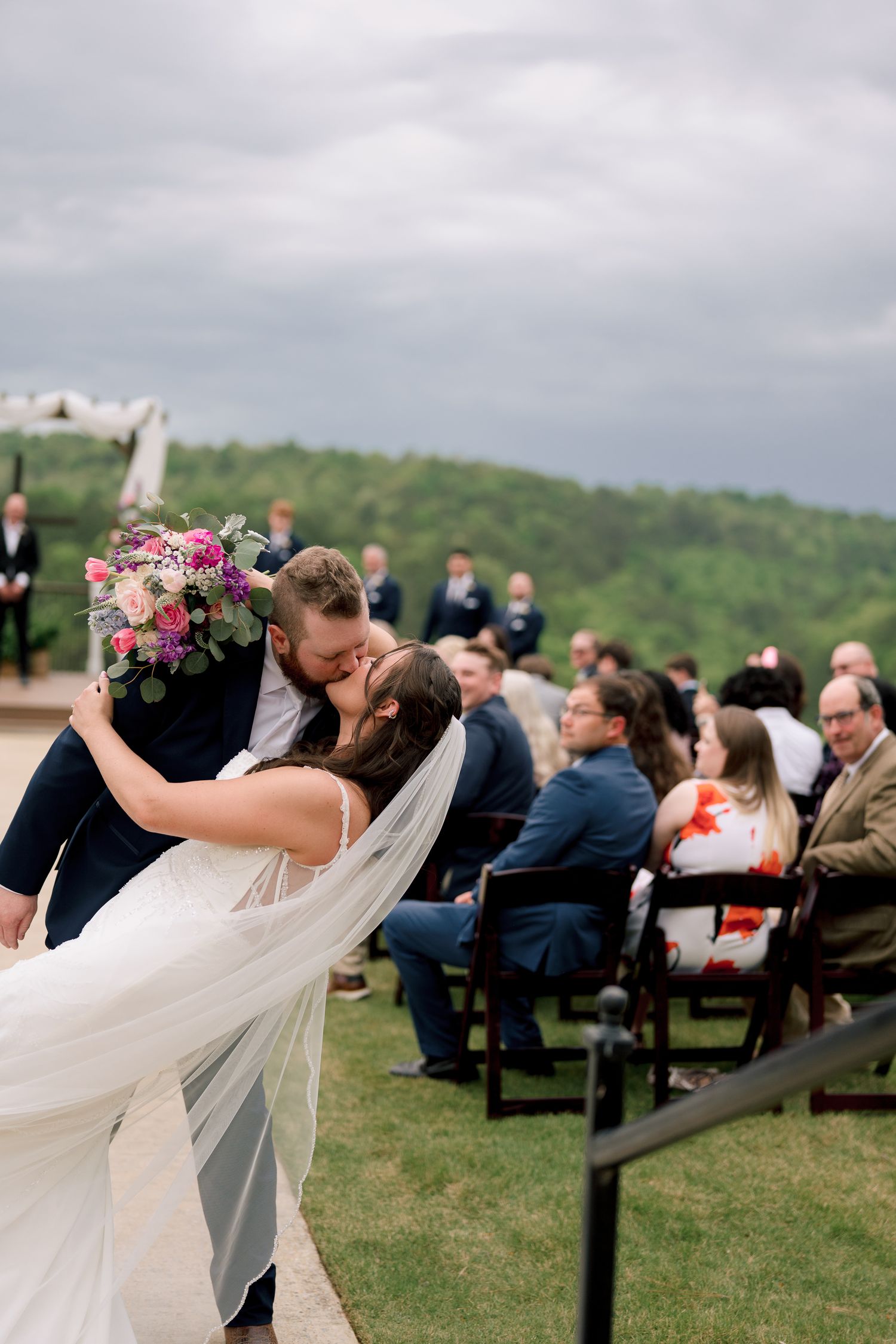 Groom dips bride for romantic kiss at outdoor wedding ceremony with seated guests and rolling hills in background.