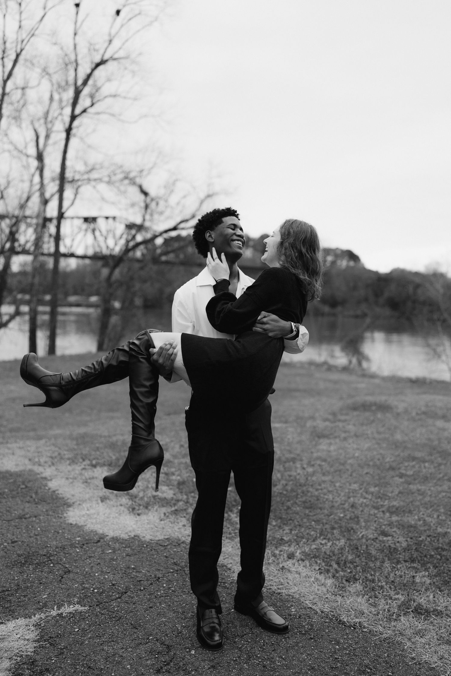 Black and white photo of a man joyfully lifting a woman in his arms near a river outdoors.