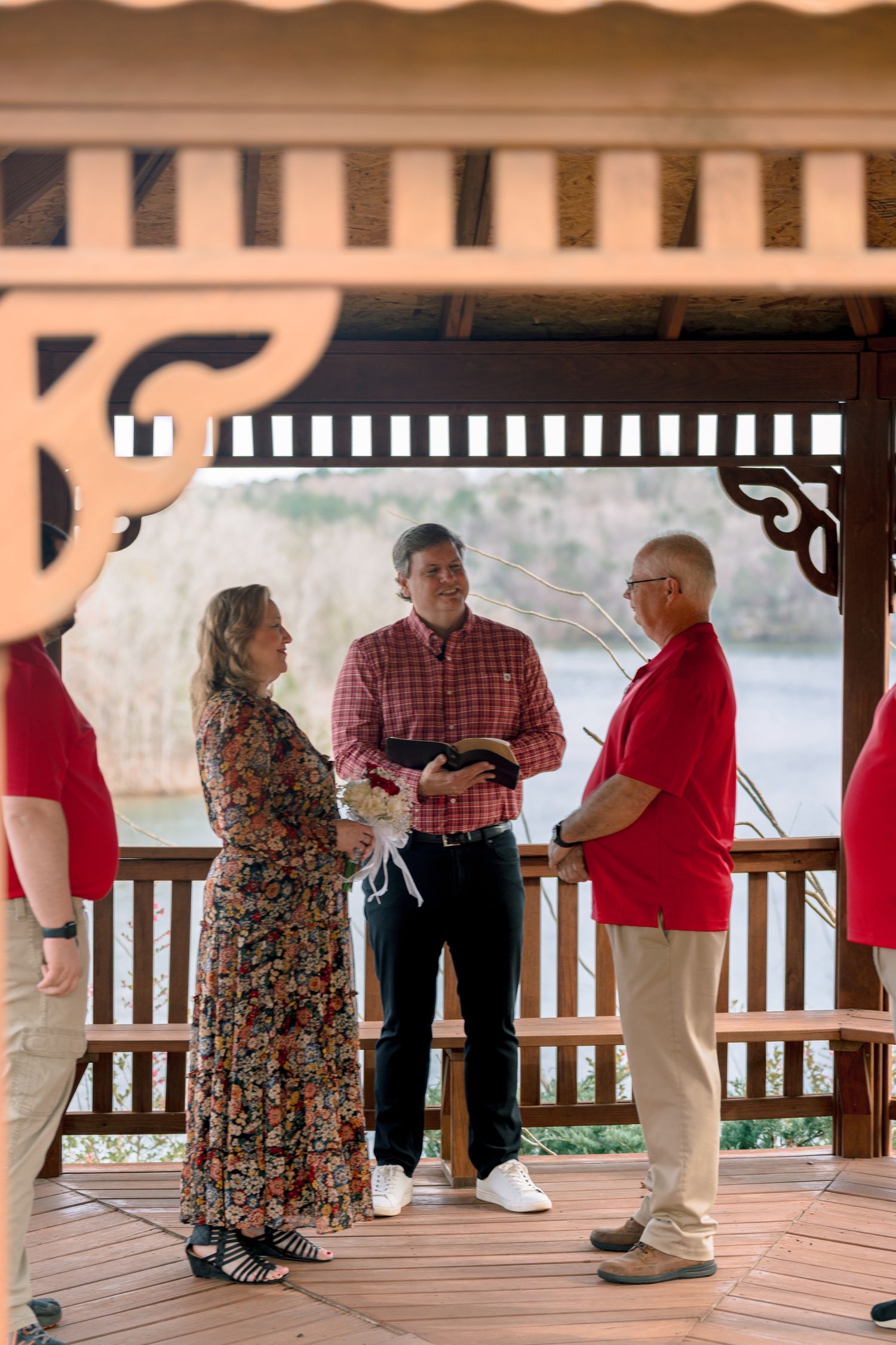 Intimate outdoor wedding ceremony in a wooden gazebo with bride in floral dress and officiant holding bible.