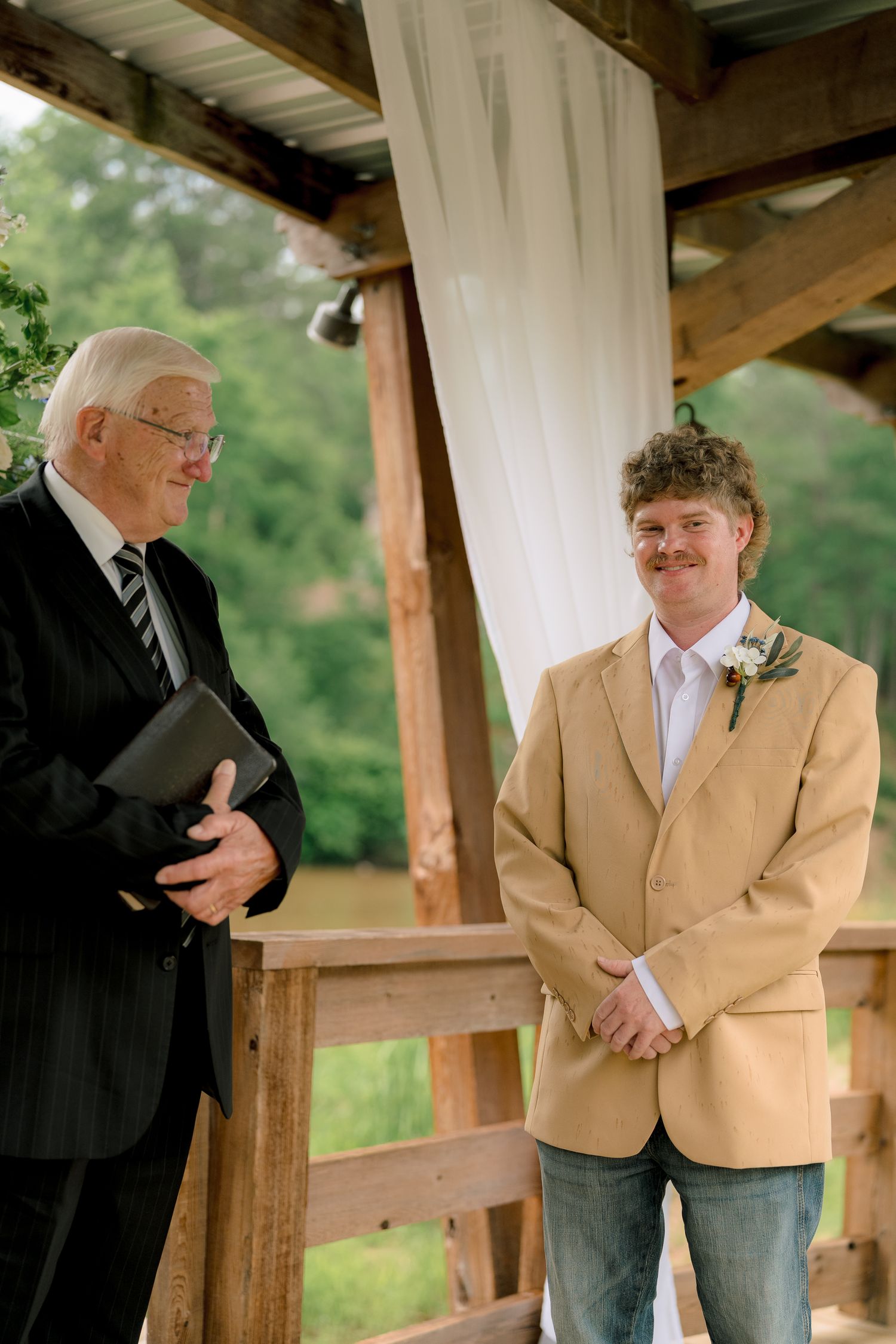 Groom in tan blazer smiles at officiant during outdoor rustic wedding ceremony under wooden pavilion.