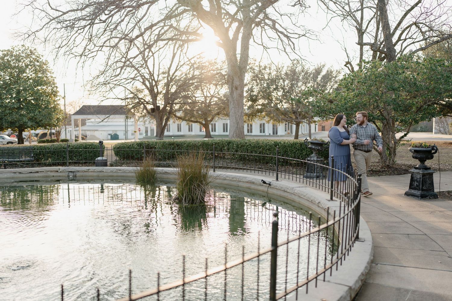 Couple standing by a decorative pond in a sunlit park with historic buildings in the background.