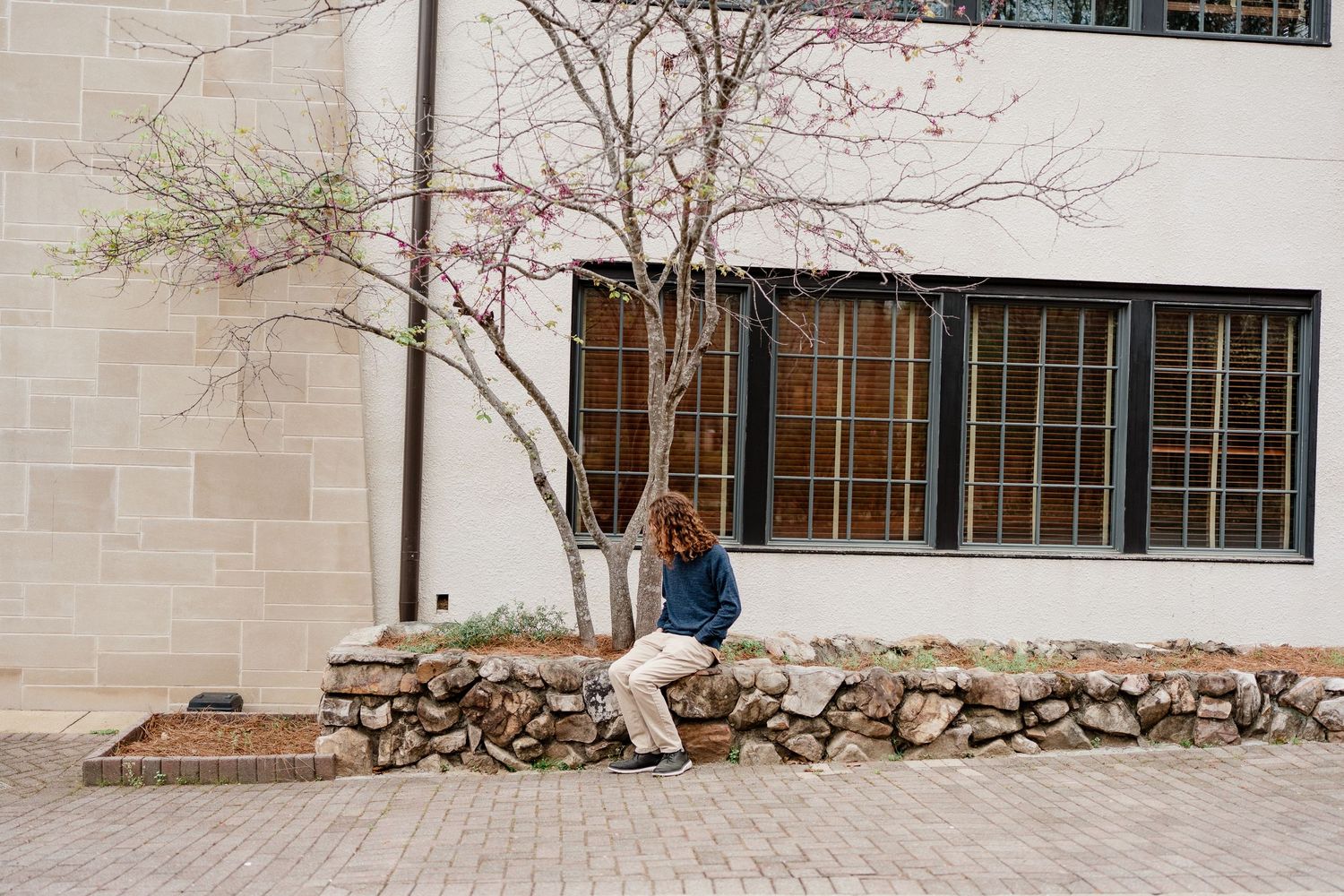 Person relaxing on stone retaining wall outside building with large windows.