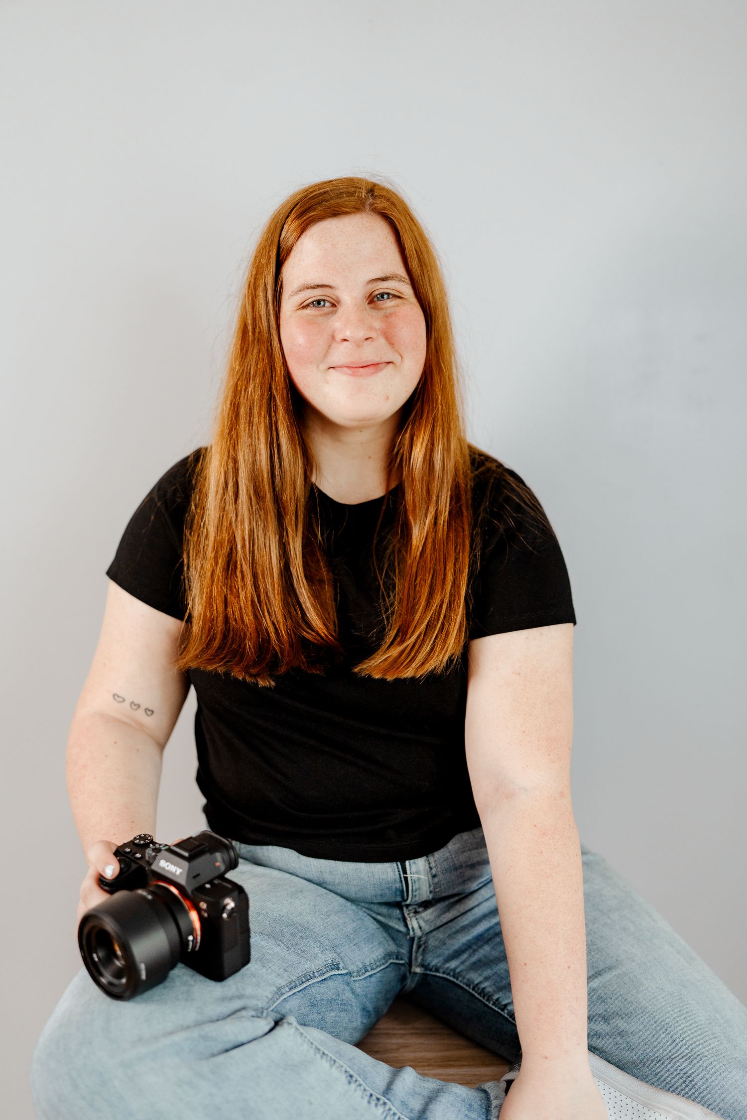 Person in casual attire poses with camera while sitting comfortably against neutral backdrop.