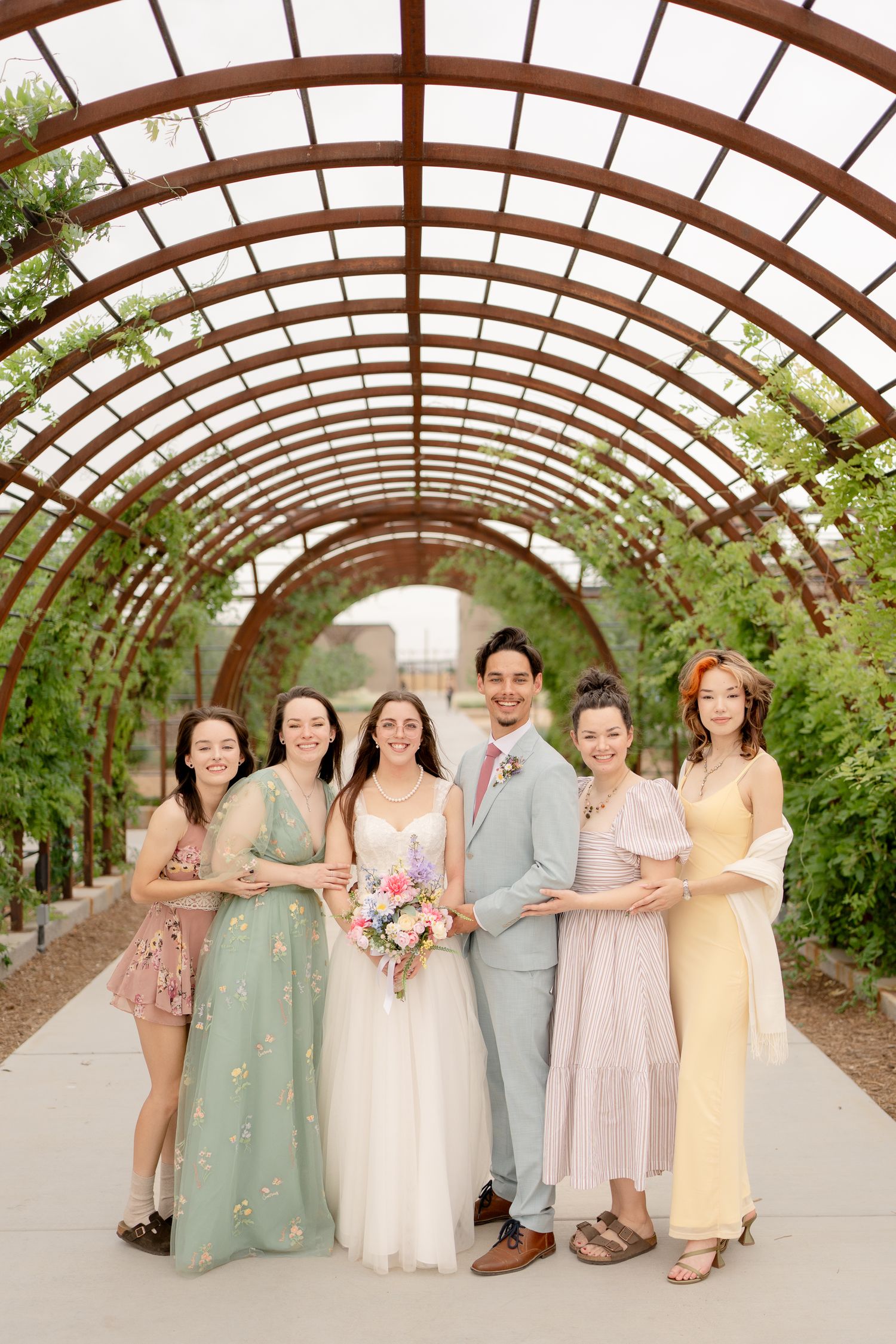 Wedding party poses together under rustic wooden arches in a greenhouse setting with soft natural light.