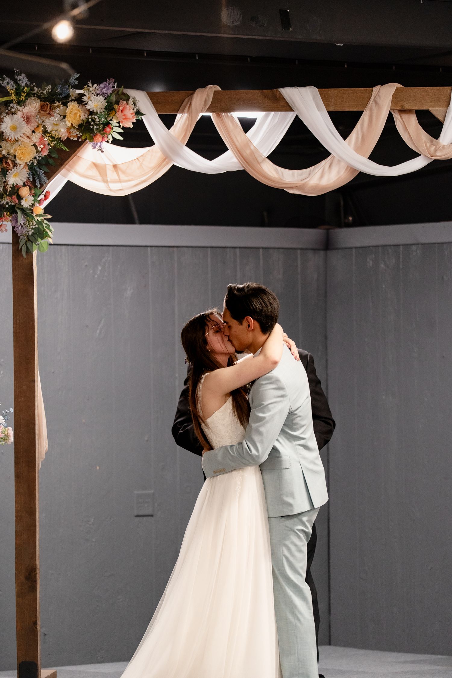 A couple shares their first kiss as newlyweds under elegant white draping and floral decor at an indoor wedding ceremony.