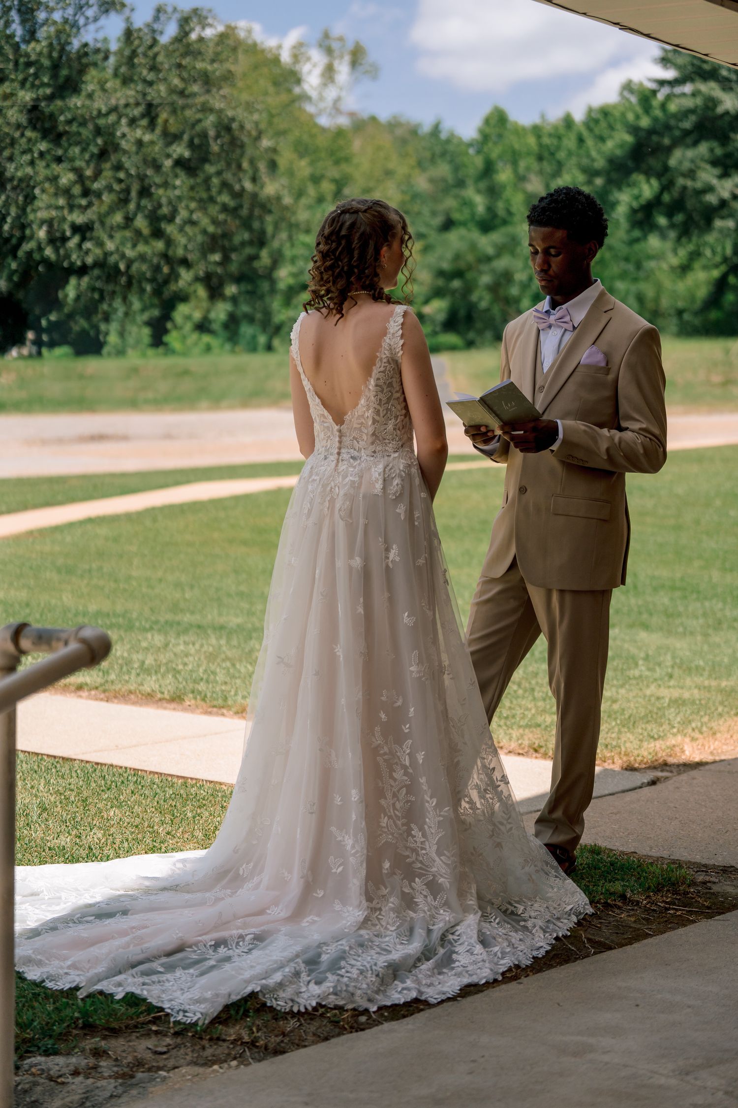 Groom in tan suit reading wedding vows to bride in lace gown outdoors in garden setting.