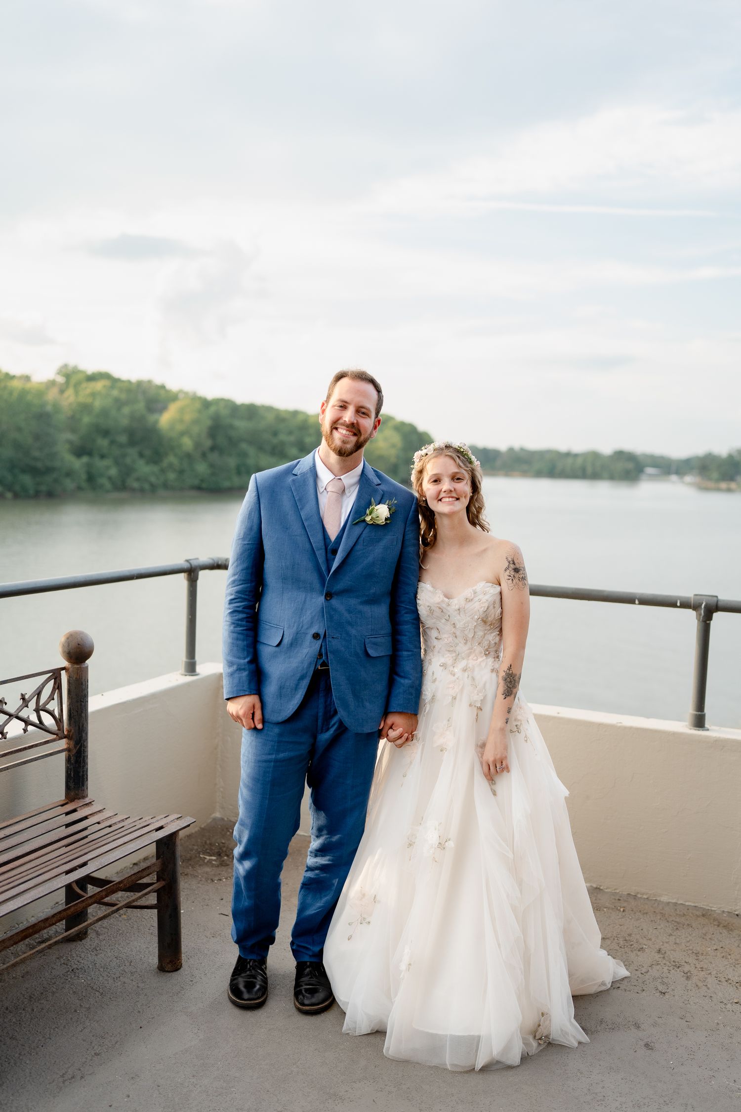 A couple poses together on a waterfront balcony in formal wedding attire.