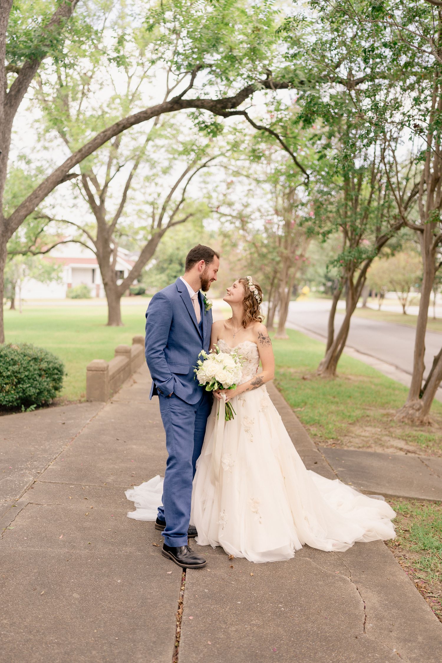 A couple in wedding attire share romantic moments along a tree-lined path in a park setting.